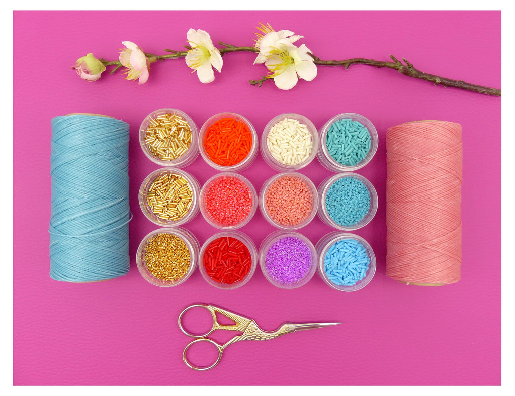Peut inclure: Assortiment de perles colorées dans de petits contenants, deux bobines de fil, une bleue et une rose, et une paire de ciseaux dorés sur un fond rose.