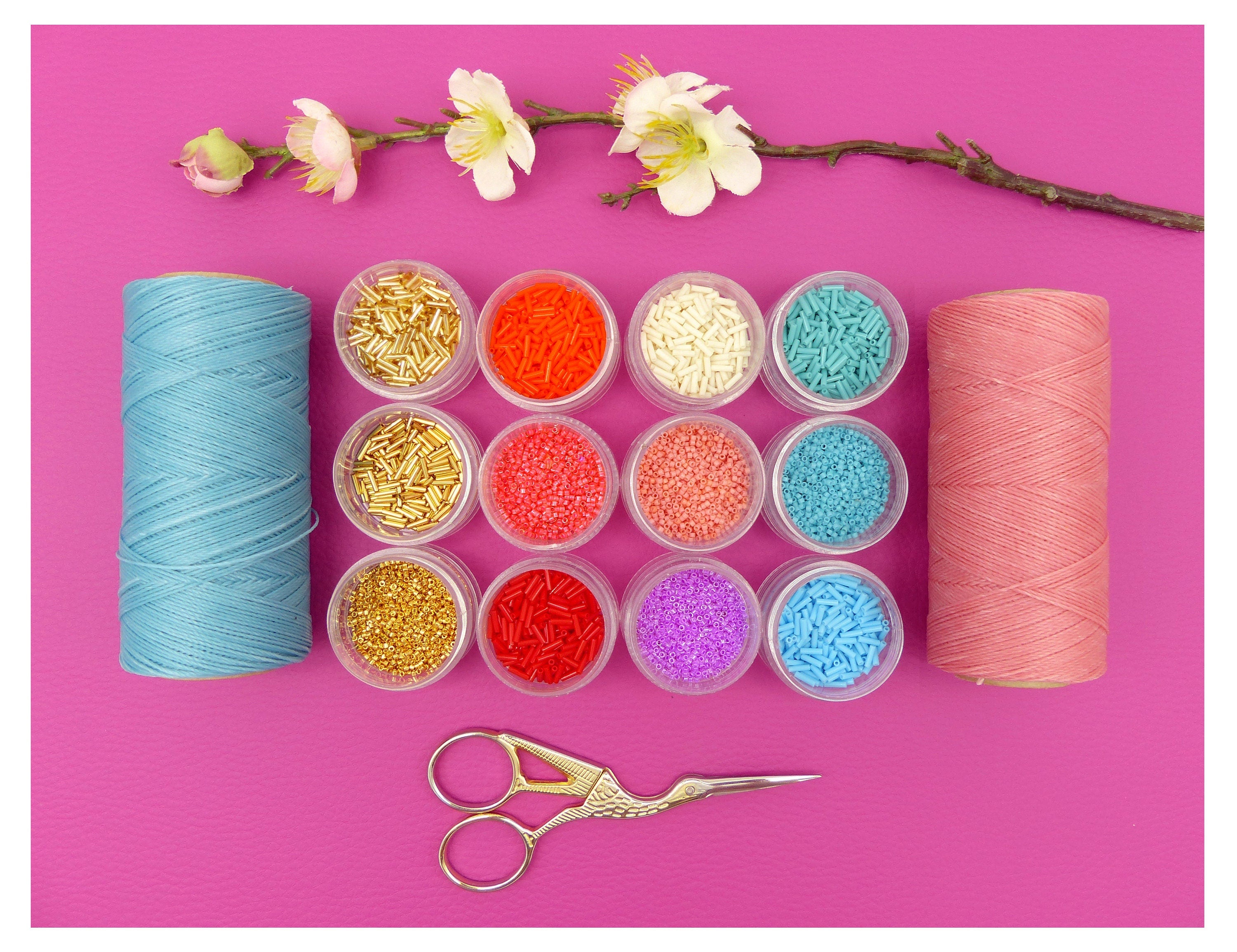 Peut inclure: Assortiment de perles colorées dans de petits contenants, deux bobines de fil, une bleue et une rose, et une paire de ciseaux dorés sur un fond rose.