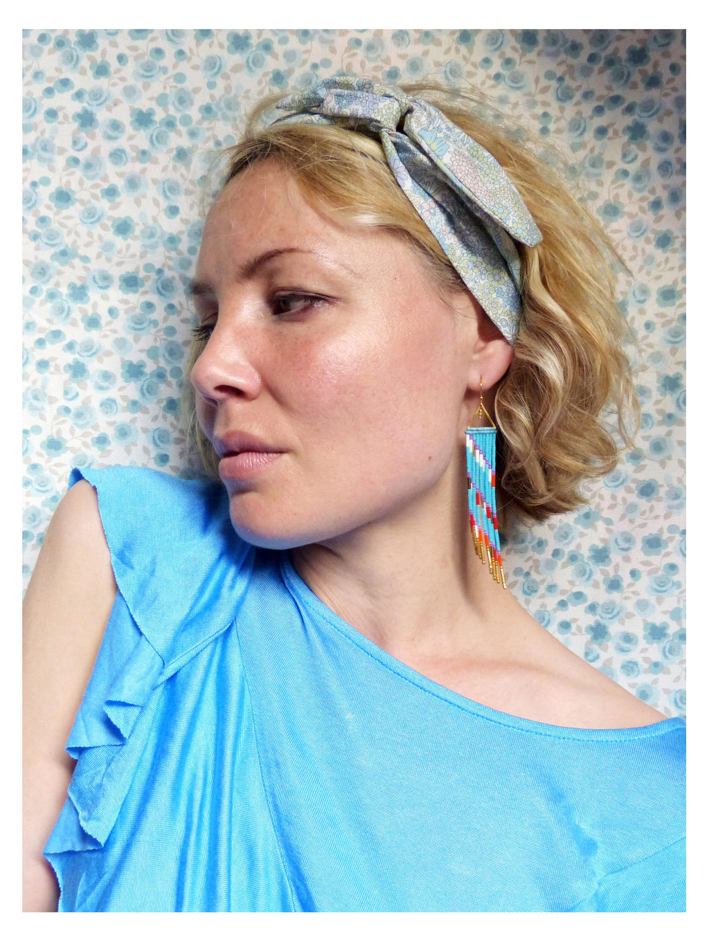 Peut inclure: Une femme aux cheveux blonds porte un haut bleu clair à volants et un bandeau à motif bleu clair et blanc. Elle porte de longues boucles d'oreilles en perles avec des accents turquoise, rouge et or.