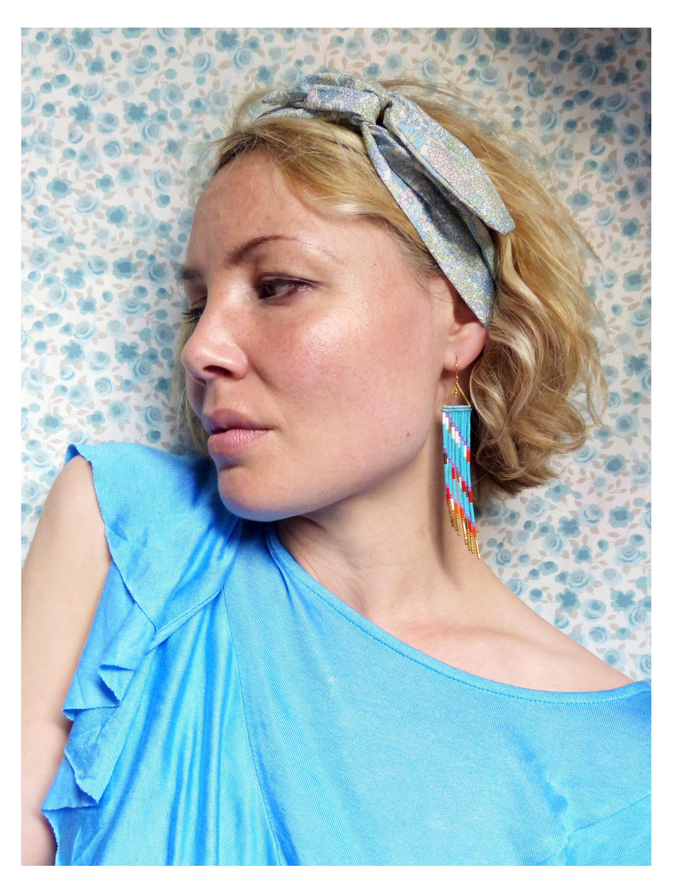 Peut inclure: Une femme aux cheveux blonds porte un haut bleu clair à volants et un bandeau à motif bleu clair et blanc. Elle porte de longues boucles d'oreilles en perles avec des accents turquoise, rouge et or.