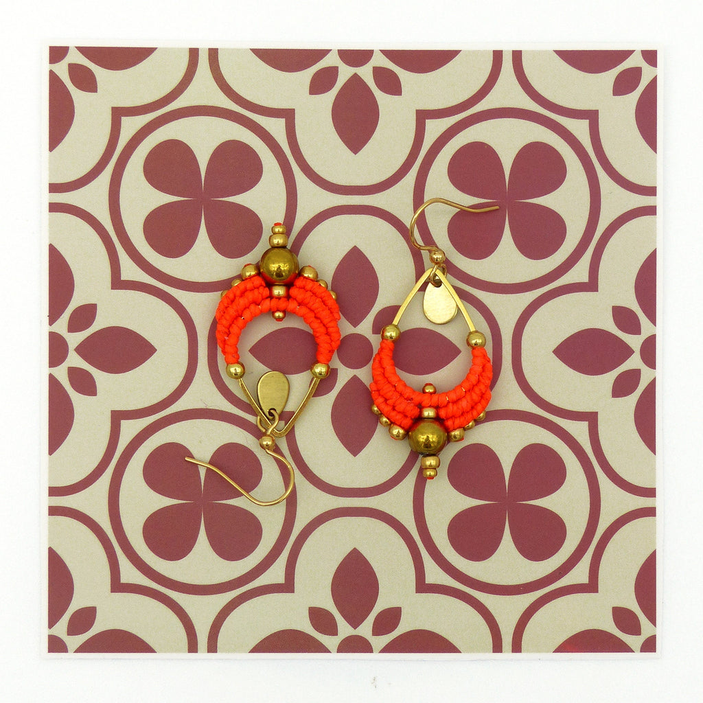 Peut inclure: Une paire de boucles d'oreilles en forme de goutte d'eau, composées de perles orange et dorées. Les boucles d'oreilles sont suspendues à un fond à motifs.