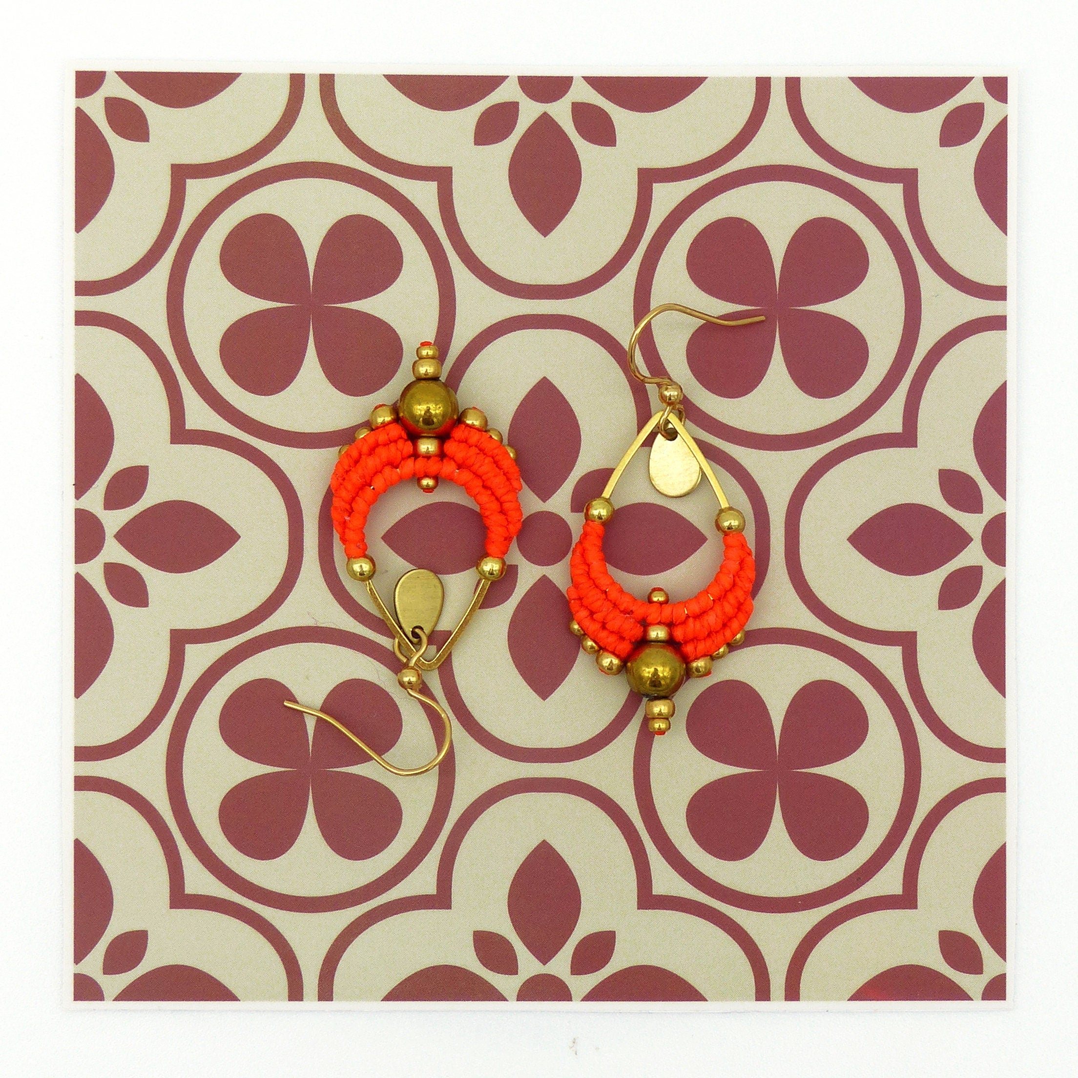 Peut inclure: Une paire de boucles d'oreilles en forme de goutte d'eau, composées de perles orange et dorées. Les boucles d'oreilles sont suspendues à un fond à motifs.