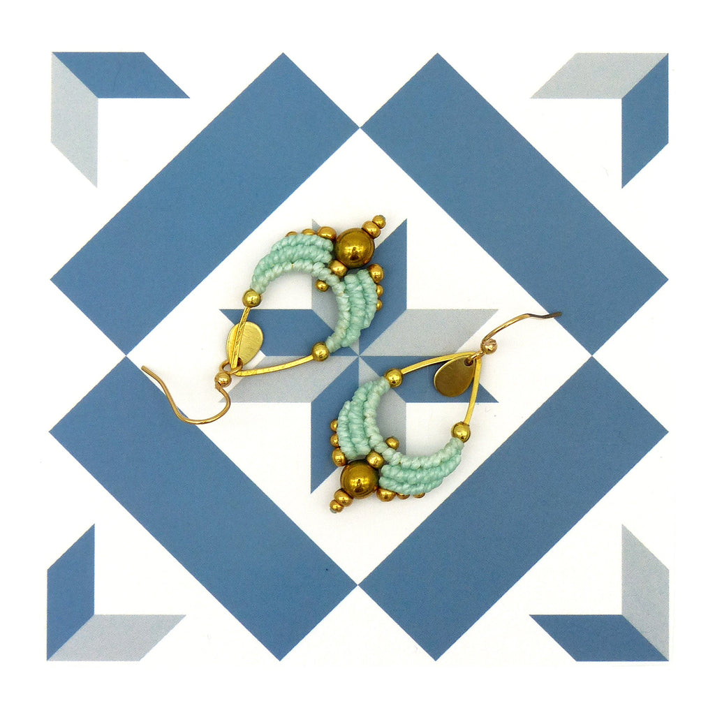 Peut inclure: Une paire de boucles d'oreilles en forme de larme, composées de perles dorées et bleu clair. Les boucles d'oreilles sont suspendues à un fond blanc avec un motif géométrique bleu et blanc.