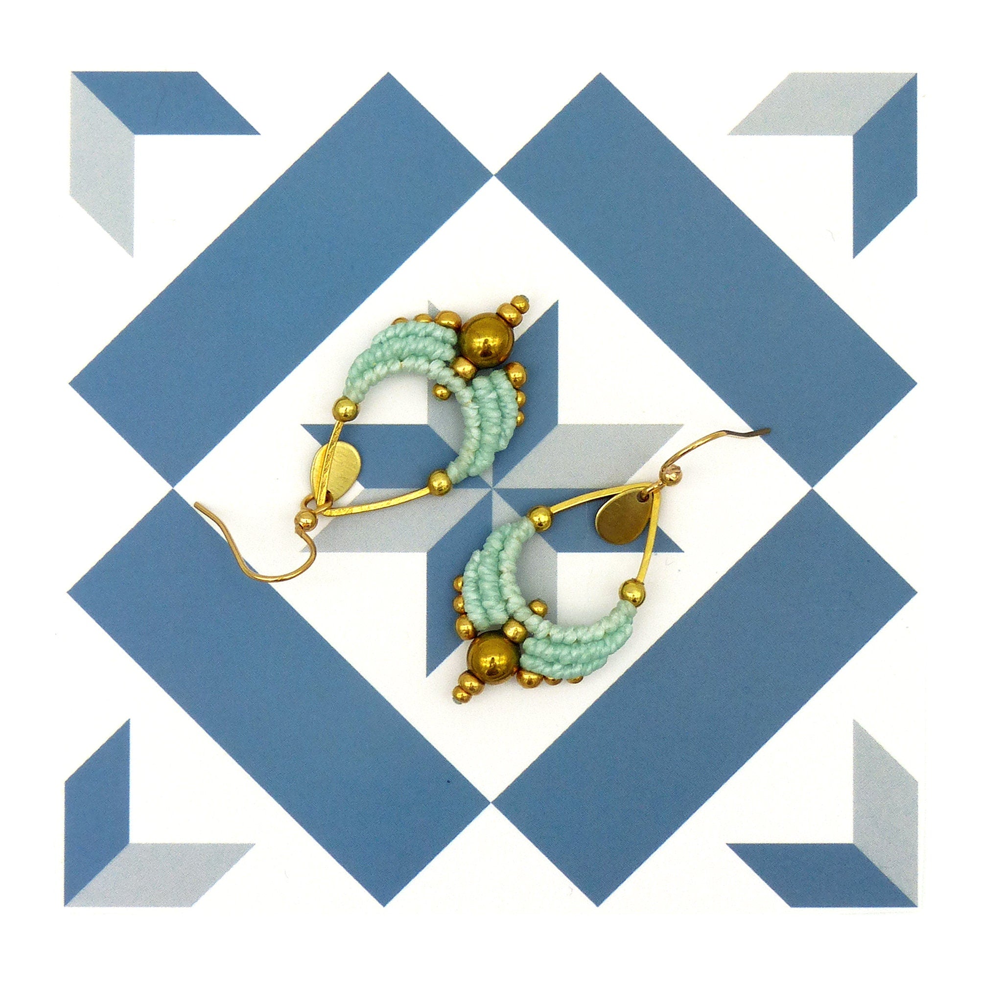 Peut inclure: Une paire de boucles d'oreilles en forme de larme, composées de perles dorées et bleu clair. Les boucles d'oreilles sont suspendues à un fond blanc avec un motif géométrique bleu et blanc.