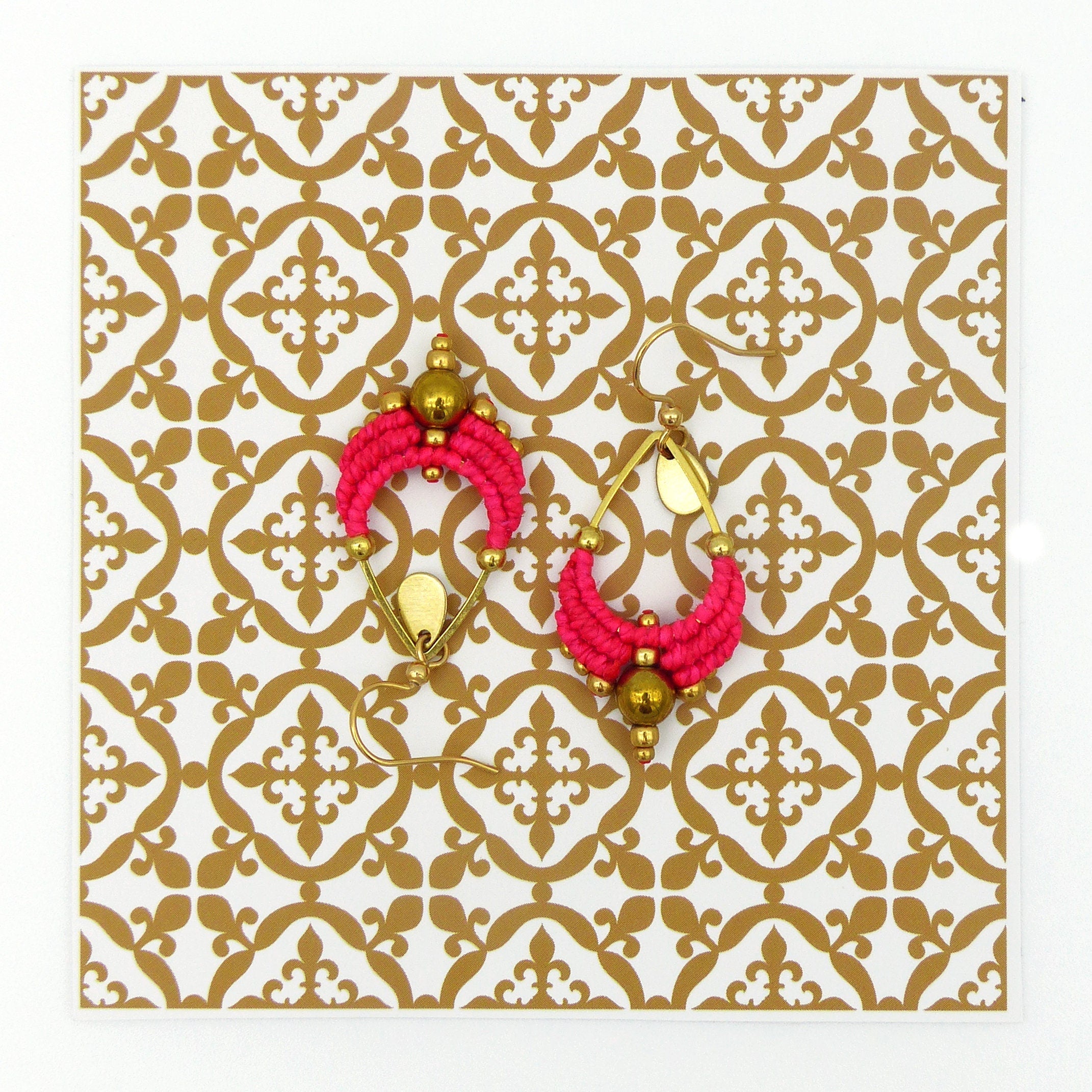 Peut inclure: Une paire de boucles d'oreilles créoles roses et dorées avec un motif perlé. Les boucles d'oreilles sont sur un fond blanc avec un motif doré.