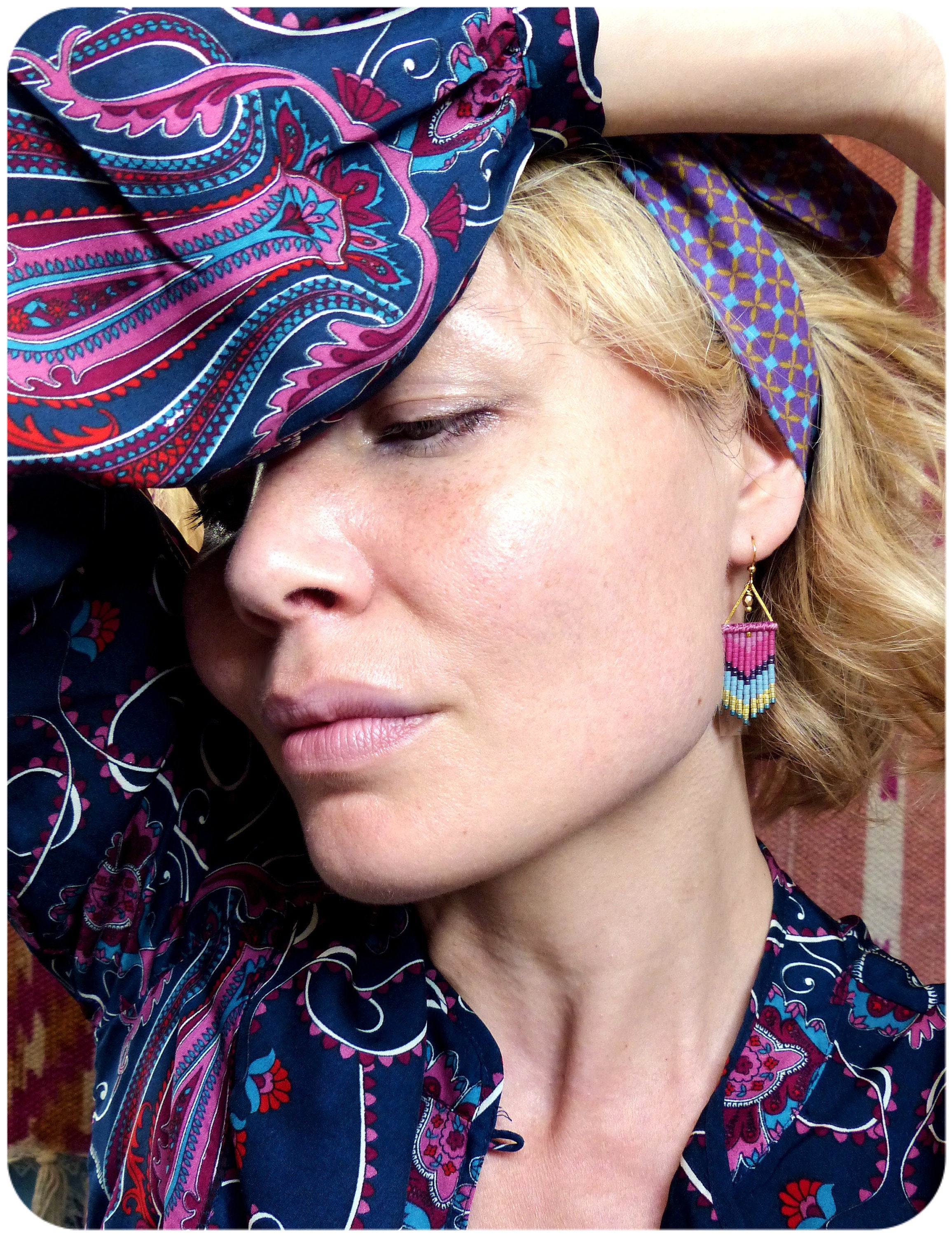 Peut inclure: Une femme portant un chemisier à motif paisley bleu et rose avec un bandeau violet et or. Elle porte des boucles d'oreilles dorées avec des franges roses et bleues.