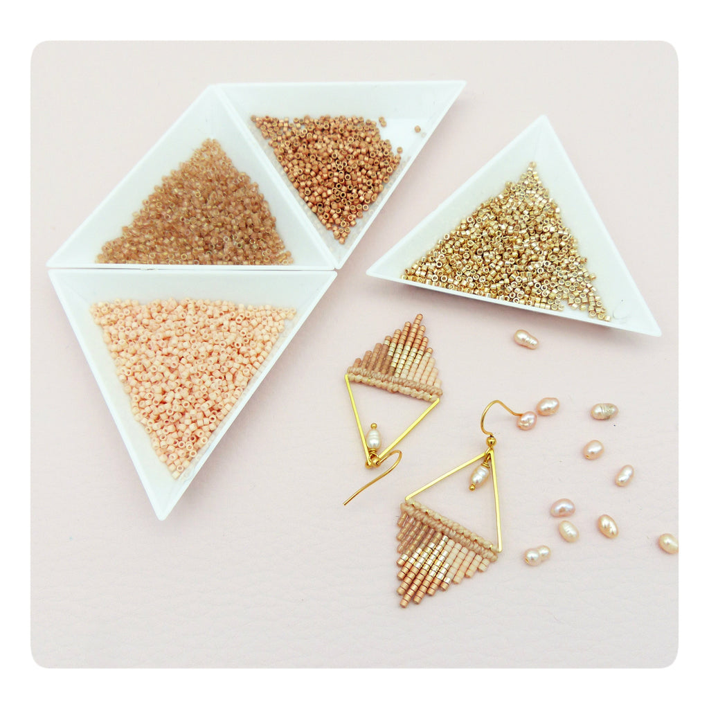 Peut inclure: Une paire de boucles d'oreilles triangulaires dorées avec des perles pêche et dorées. Les boucles d'oreilles sont présentées sur une surface rose avec de petites perles blanches et trois bols de perles en pêche, or et brun.