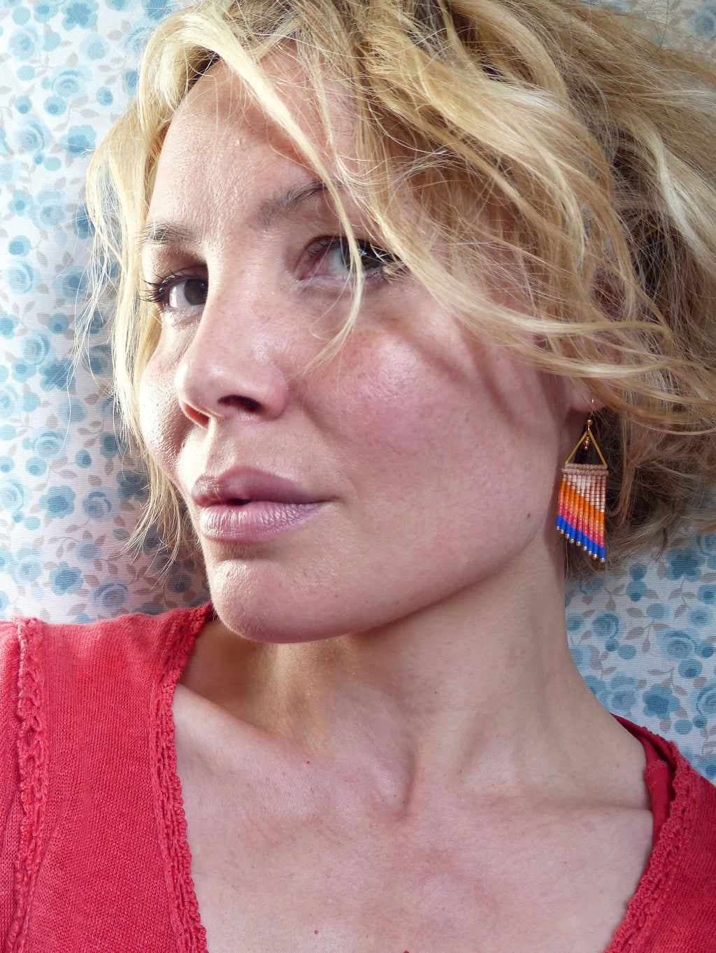 Peut inclure: Gros plan d'une personne portant un haut rouge et des boucles d'oreilles en perles. Les boucles d'oreilles ont un dessus triangulaire avec une frange de perles orange, bleues et blanches. Le fond est un motif floral bleu clair.