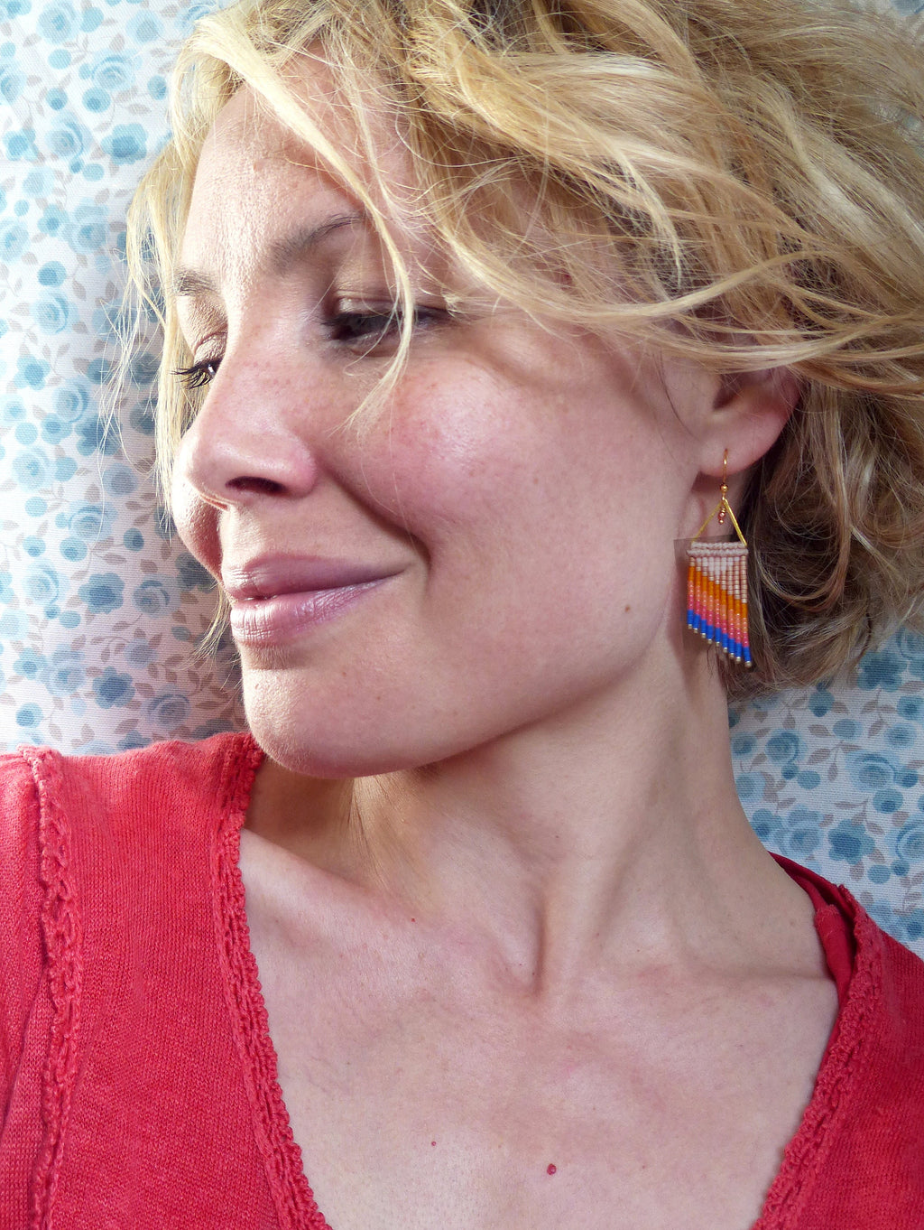 Peut inclure: Une femme porte des boucles d'oreilles en perles colorées. Les boucles d'oreilles rectangulaires ont un triangle doré en haut et un motif de perles orange, roses, blanches et bleues. Elle porte un haut rouge.