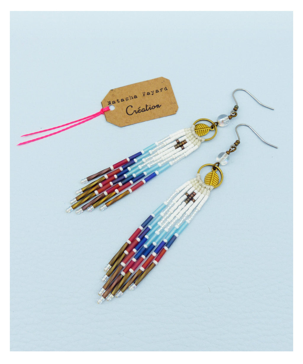 Peut inclure: Une paire de longues boucles d'oreilles en perles avec un motif blanc, bleu, rouge et marron. Les boucles d'oreilles ont un petit charme en forme de feuille en haut et sont suspendues à un crochet argenté.