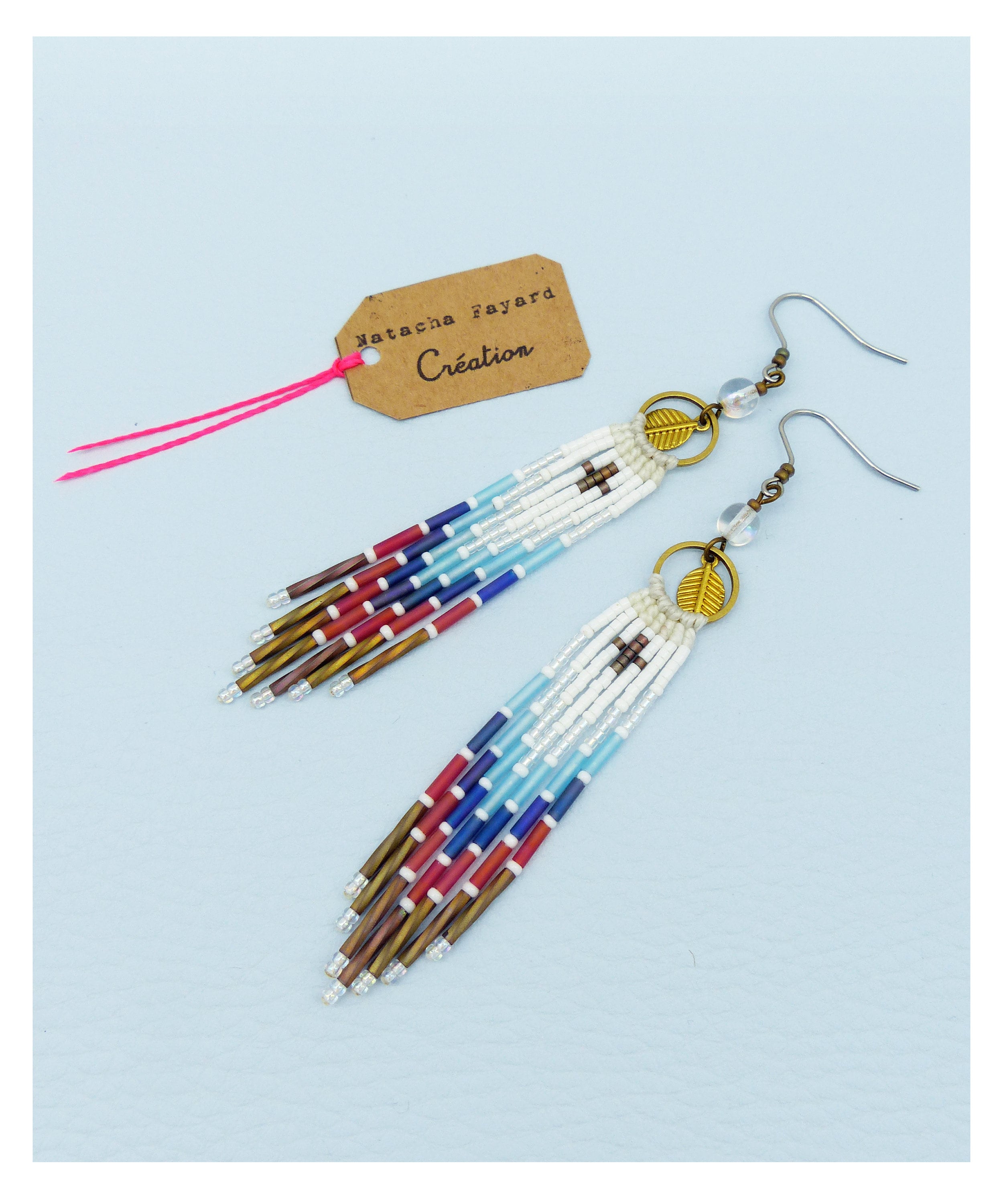 Peut inclure: Une paire de longues boucles d'oreilles en perles avec un motif blanc, bleu, rouge et marron. Les boucles d'oreilles ont un petit charme en forme de feuille en haut et sont suspendues à un crochet argenté.
