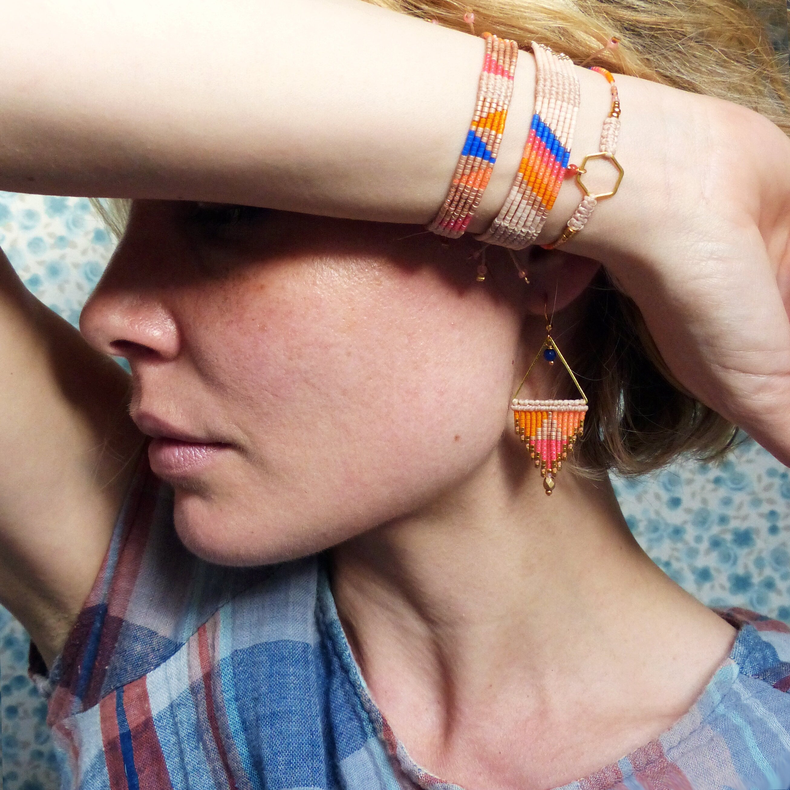 Peut inclure: Une femme portant une chemise à carreaux bleus et rouges avec une paire de boucles d'oreilles en perles. Les boucles d'oreilles sont en or avec un motif géométrique et comportent des perles orange, roses et bleues. La femme porte également plusieurs bracelets en perles au poignet.