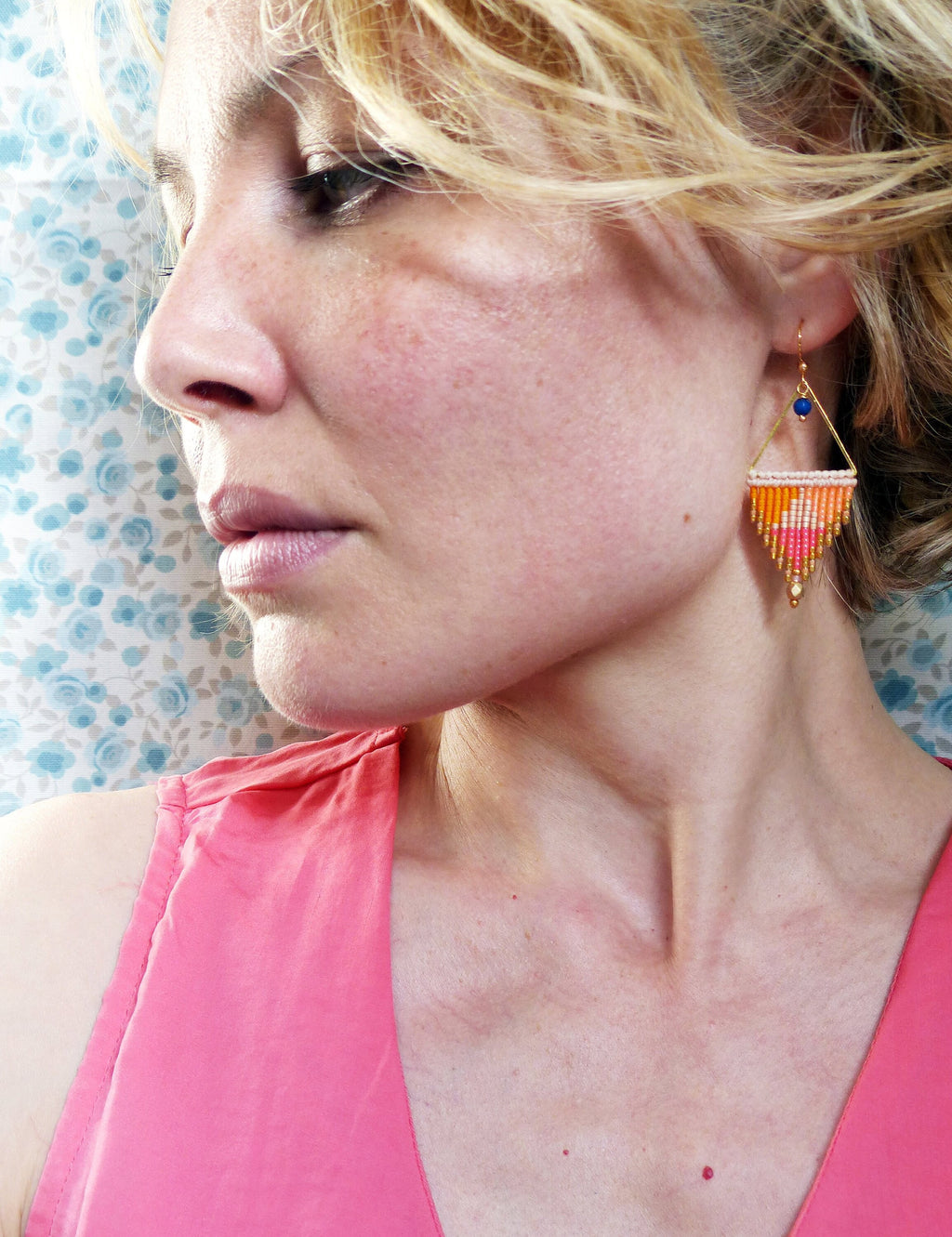 Peut inclure: Une femme portant un haut rose sans manches avec une paire de boucles d'oreilles en perles dorées et orange. Les boucles d'oreilles sont de forme triangulaire et ont une petite perle bleue au sommet.