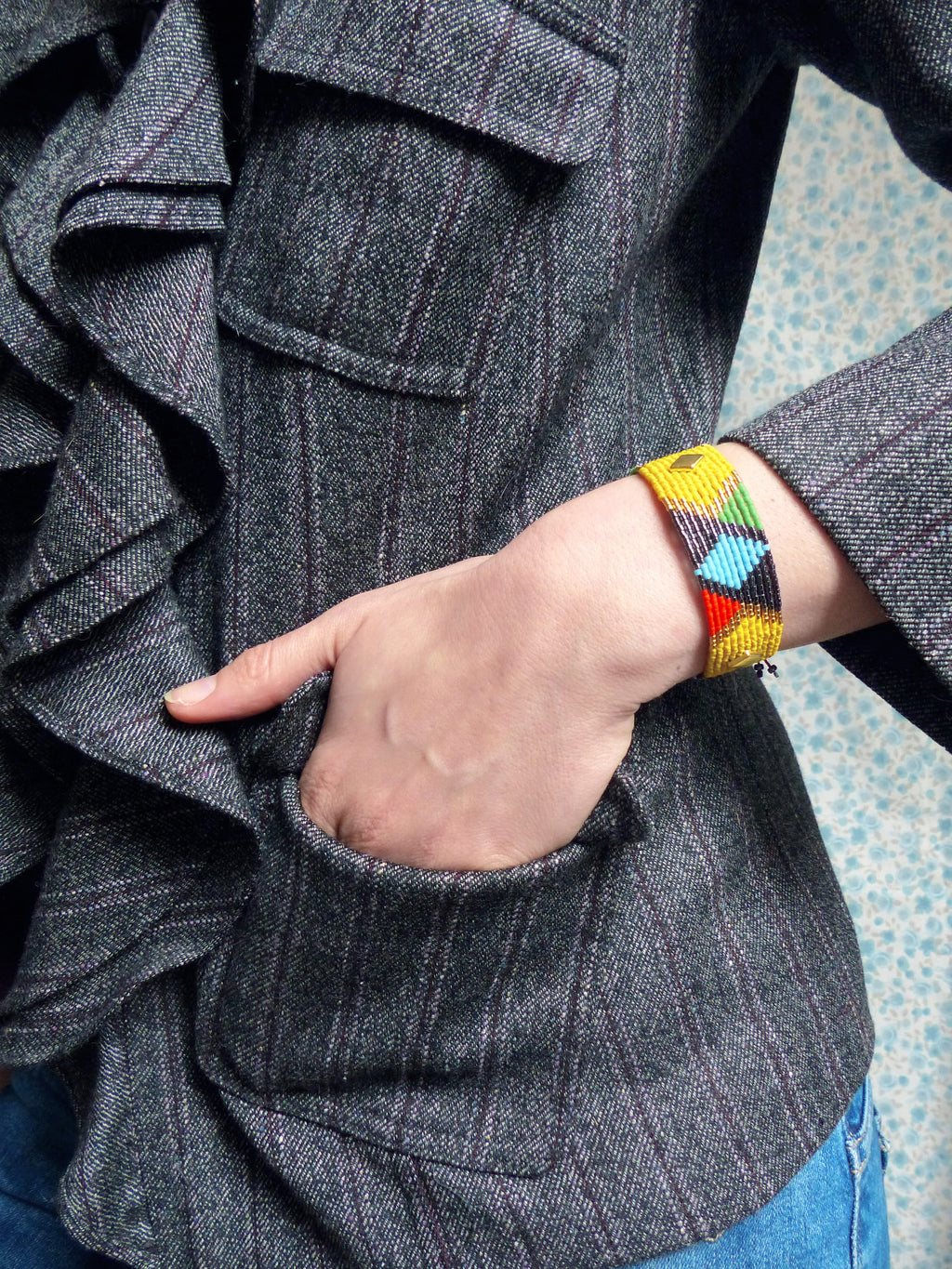 Peut inclure: Une veste en tweed gris avec un col à volants et une poche. La veste a un bracelet en perles avec des perles jaunes, bleues, rouges et noires.