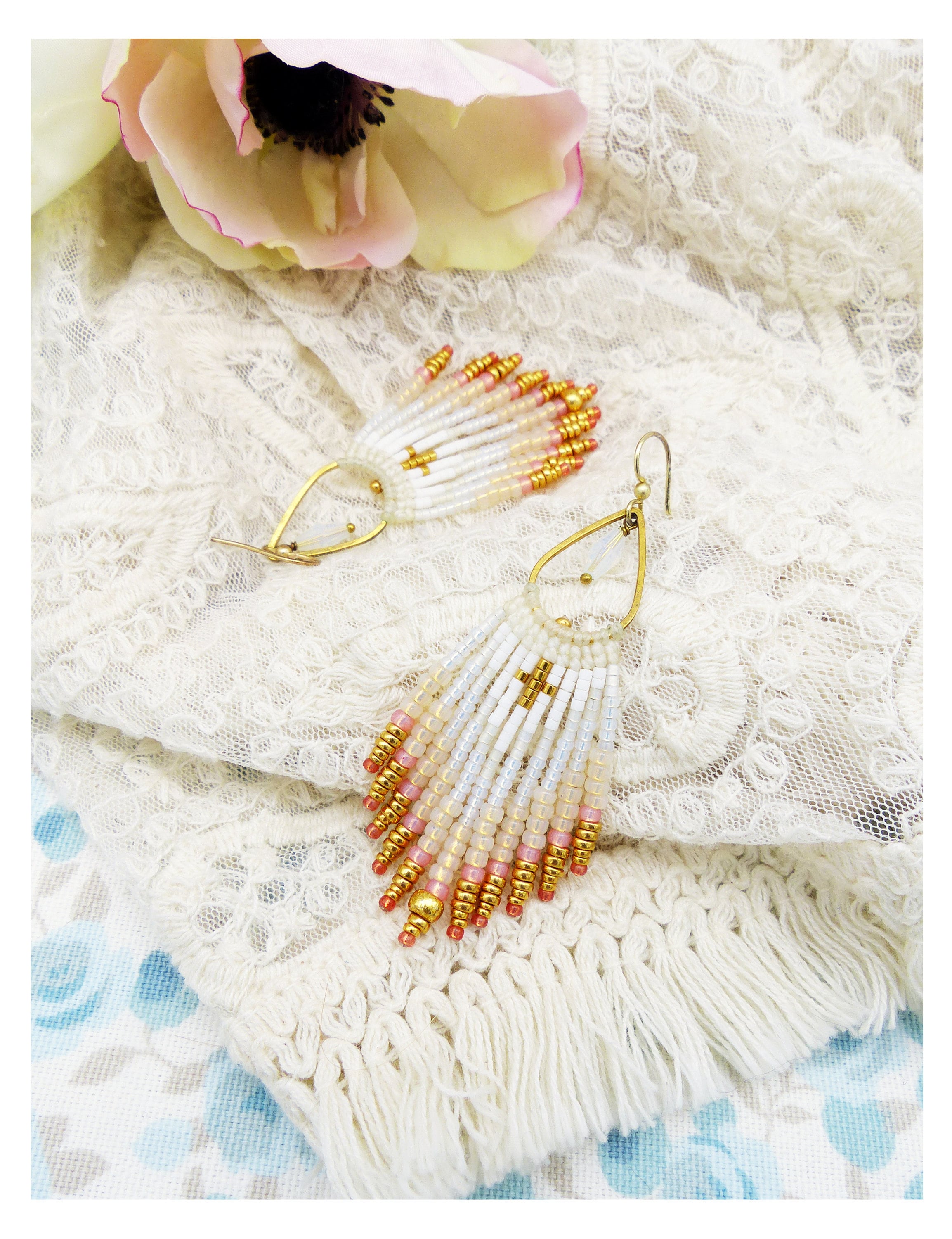 Peut inclure: Une paire de boucles d'oreilles en perles dorées et blanches avec une forme de larme et une frange de perles. Les boucles d'oreilles sont sur un fond de dentelle blanche.