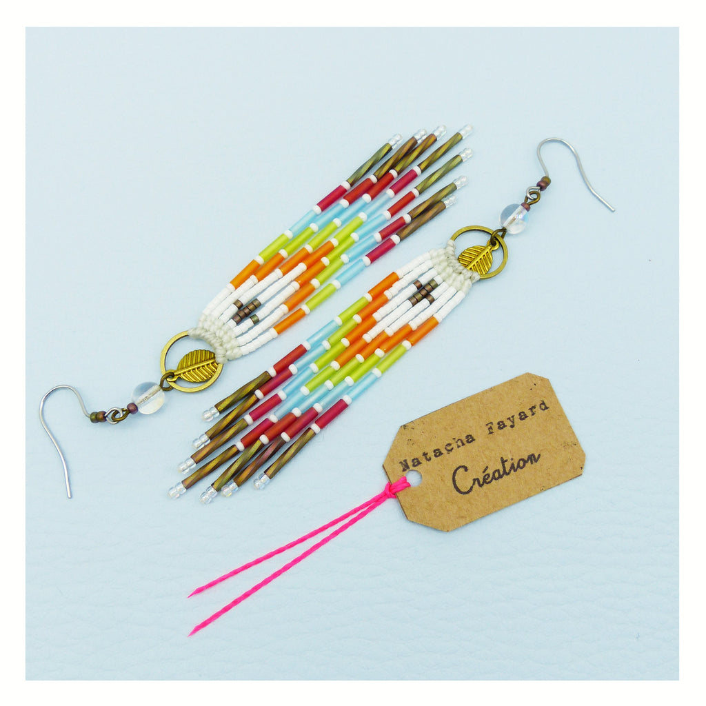 Peut inclure: Une paire de longues boucles d'oreilles en perles colorées avec un charme en feuille d'or. Les boucles d'oreilles sont faites de perles blanches, rouges, orange, jaunes, vertes et bleues. Les boucles d'oreilles sont suspendues à des crochets argentés. Les boucles d'oreilles sont sur un fond blanc avec une ficelle rose et une étiquette brune qui dit "Natasha Fayard Création".