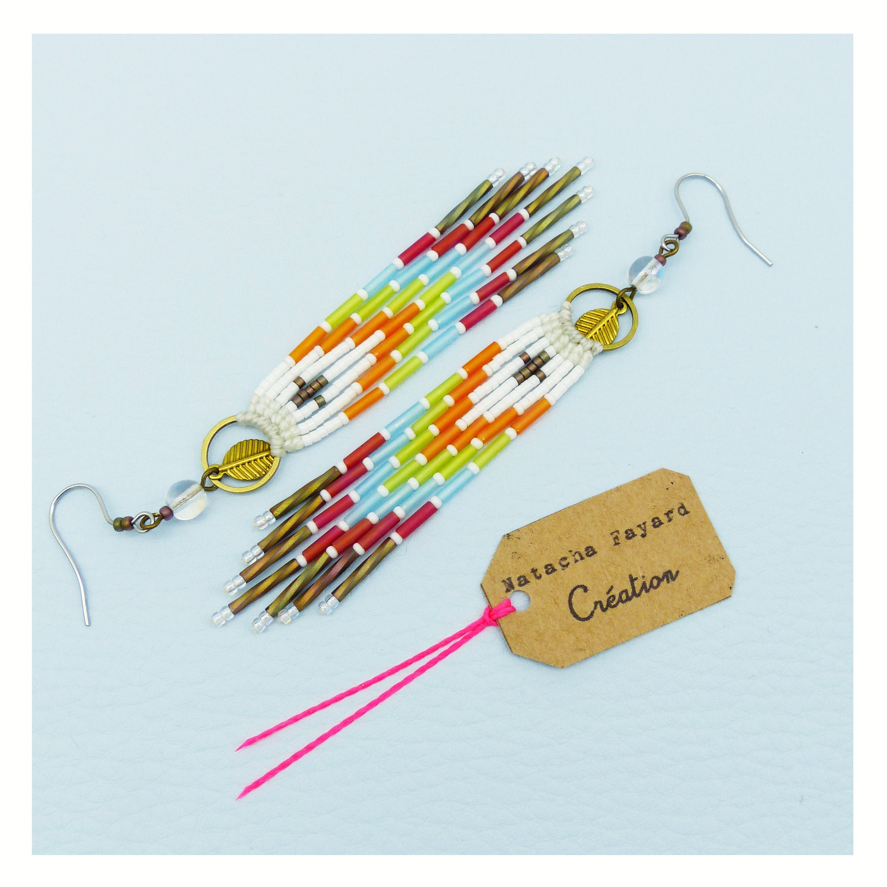 Peut inclure: Une paire de longues boucles d'oreilles en perles colorées avec un charme en feuille d'or. Les boucles d'oreilles sont faites de perles blanches, rouges, orange, jaunes, vertes et bleues. Les boucles d'oreilles sont suspendues à des crochets argentés. Les boucles d'oreilles sont sur un fond blanc avec une ficelle rose et une étiquette brune qui dit "Natasha Fayard Création".
