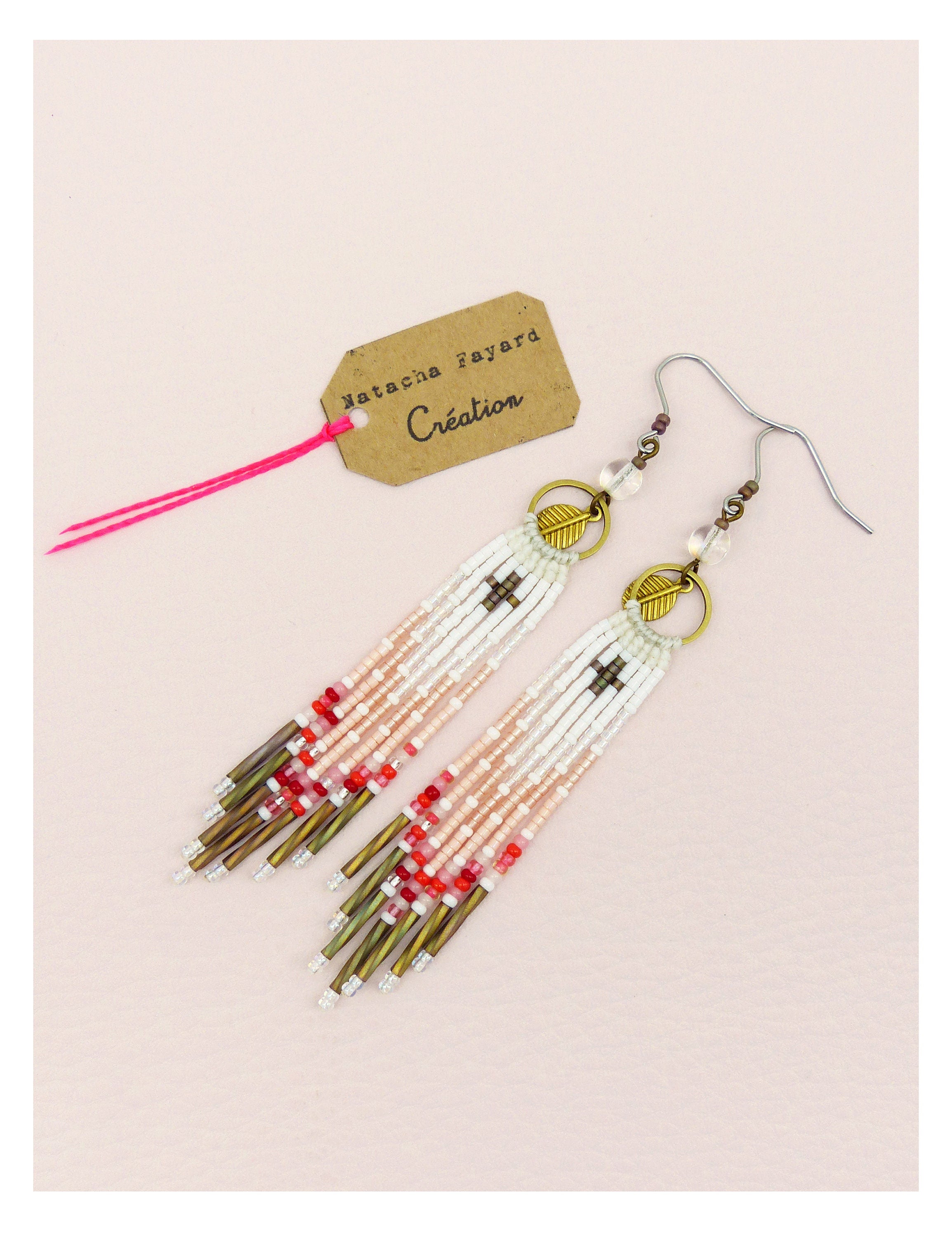 Peut inclure: Boucles d'oreilles faites à la main avec un design à longues franges. Les boucles d'oreilles sont composées de perles blanches, roses, rouges et bronze, avec un motif de croix en haut. Elles portent l'inscription "Natacha Fayard Création".