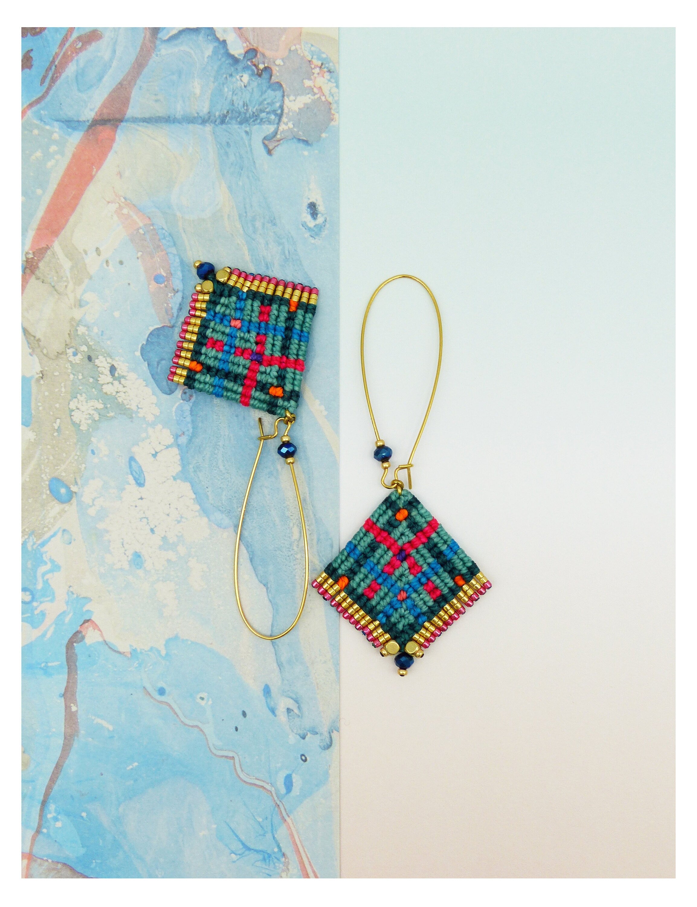 Peut inclure: Une paire de boucles d'oreilles macramé faites à la main. Elles présentent un motif carré turquoise, rouge et orange, agrémenté de perles dorées et d'un fil doré. Une boucle d'oreille est détachée, révélant le design complet.