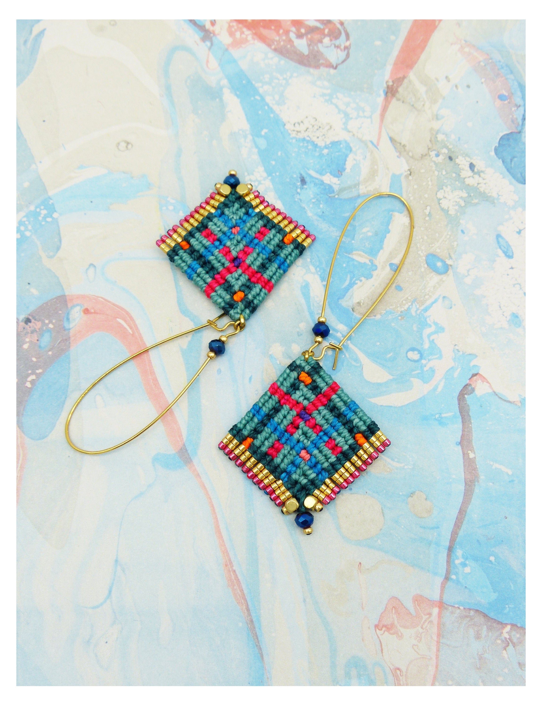 Peut inclure: Une paire de boucles d'oreilles macramé faites à la main. Chaque boucle d'oreille présente un motif en losange en bleu sarcelle, rose et orange, orné de perles dorées et bleues. Les boucles d'oreilles sont suspendues à de longs crochets d'oreilles dorés.