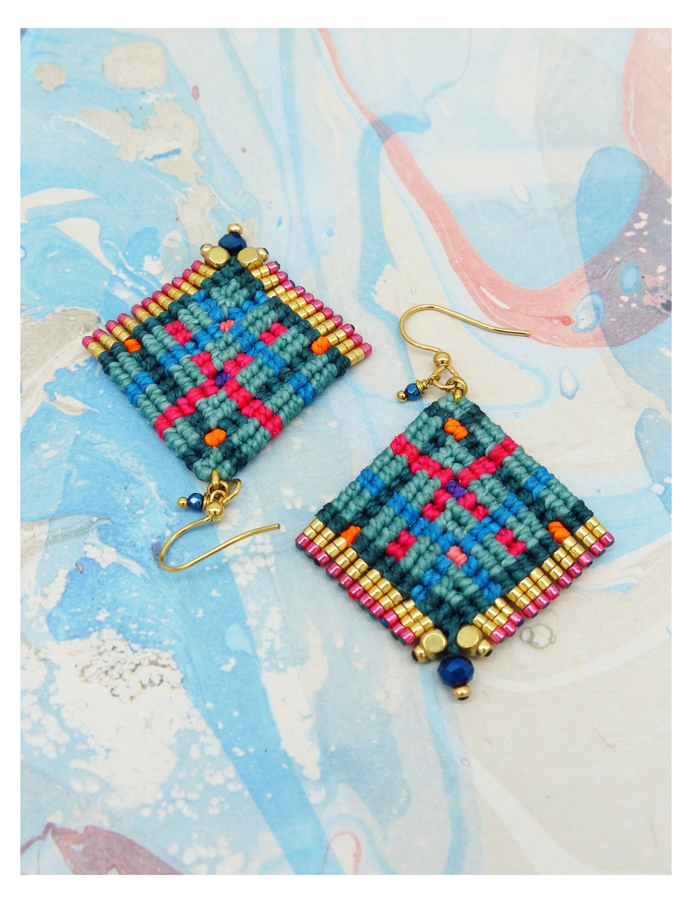 Peut inclure: Paire de boucles d'oreilles macramé faites à la main. Chaque boucle d'oreille présente un motif en forme de losange avec des fils tissés turquoise, roses, bleus et oranges. Les bords sont bordés de perles dorées et les boucles ont des crochets dorés.