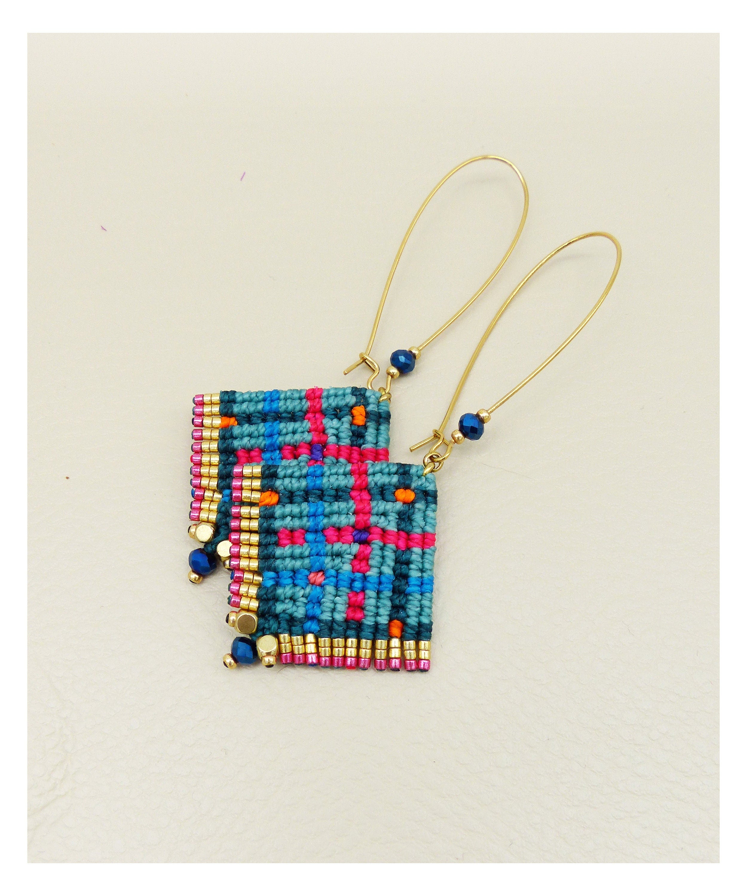 Peut inclure: Une paire de boucles d'oreilles carrées faites à la main avec un motif macramé tissé. Les boucles d'oreilles présentent un motif à carreaux bleu sarcelle, rose et orange, rehaussé de perles dorées et bleues. Les longues attaches d'oreilles dorées ajoutent une touche délicate.