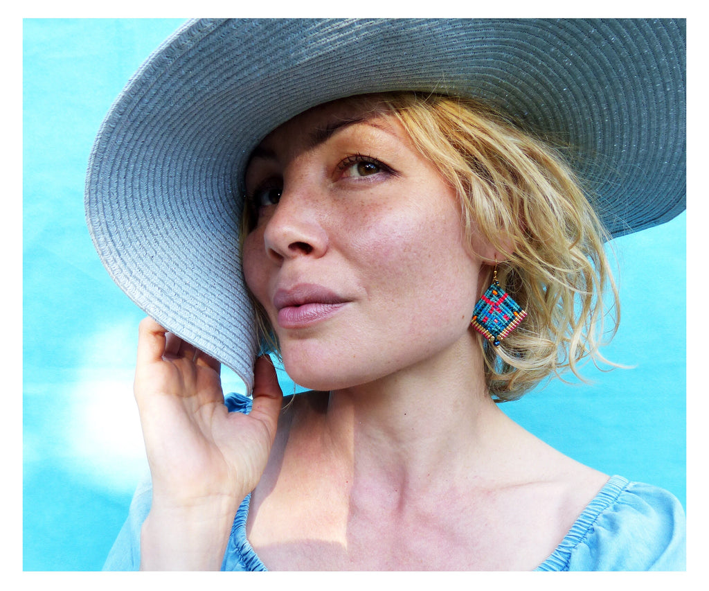 Peut inclure: Une femme porte un chapeau de soleil bleu clair et un haut bleu. Elle porte des boucles d'oreilles géométriques colorées avec des accents rouges, bleus et dorés. Le fond est bleu vif et uni.