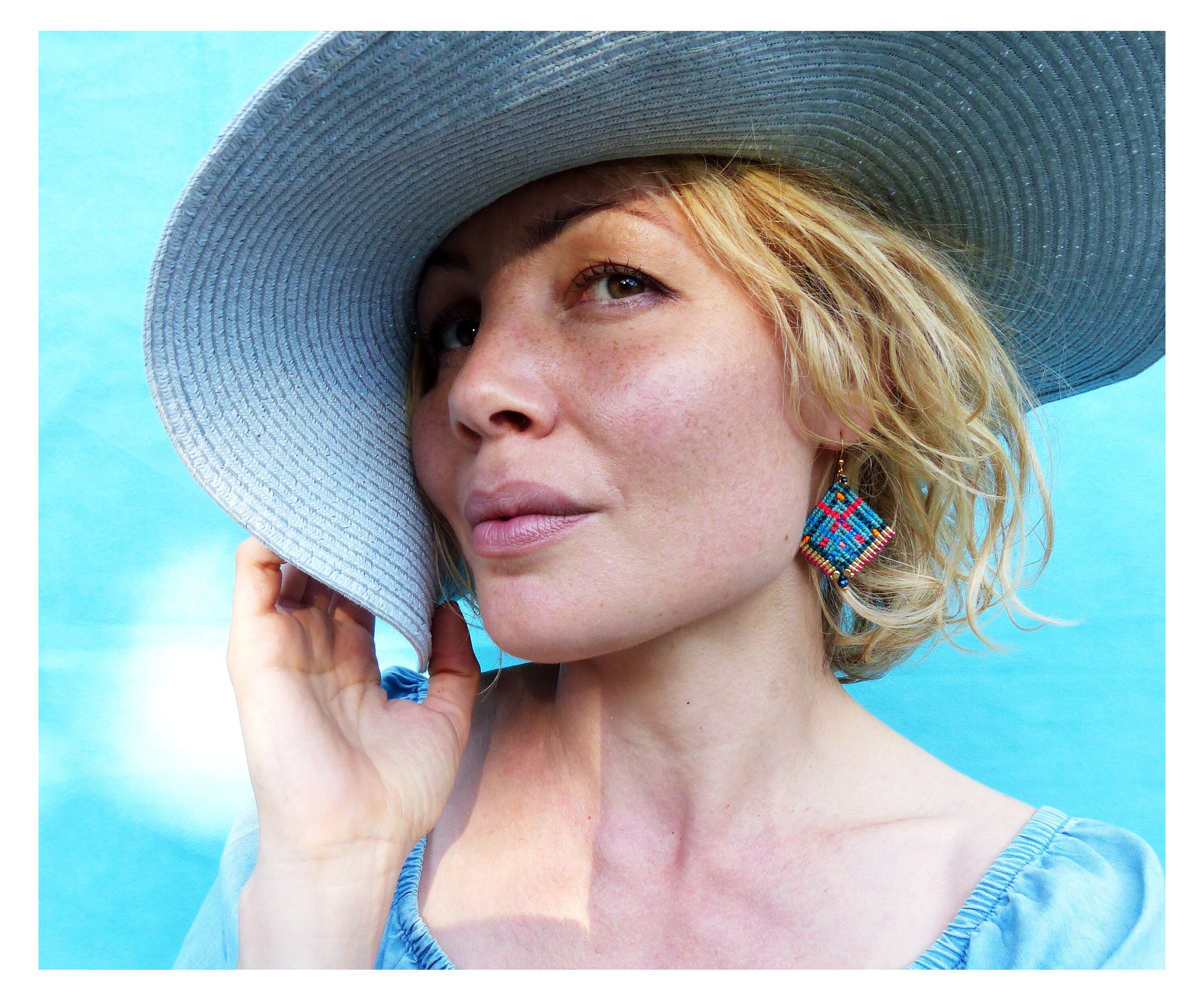 Peut inclure: Une femme porte un chapeau de soleil bleu clair et un haut bleu. Elle porte des boucles d'oreilles géométriques colorées avec des accents rouges, bleus et dorés. Le fond est bleu vif et uni.