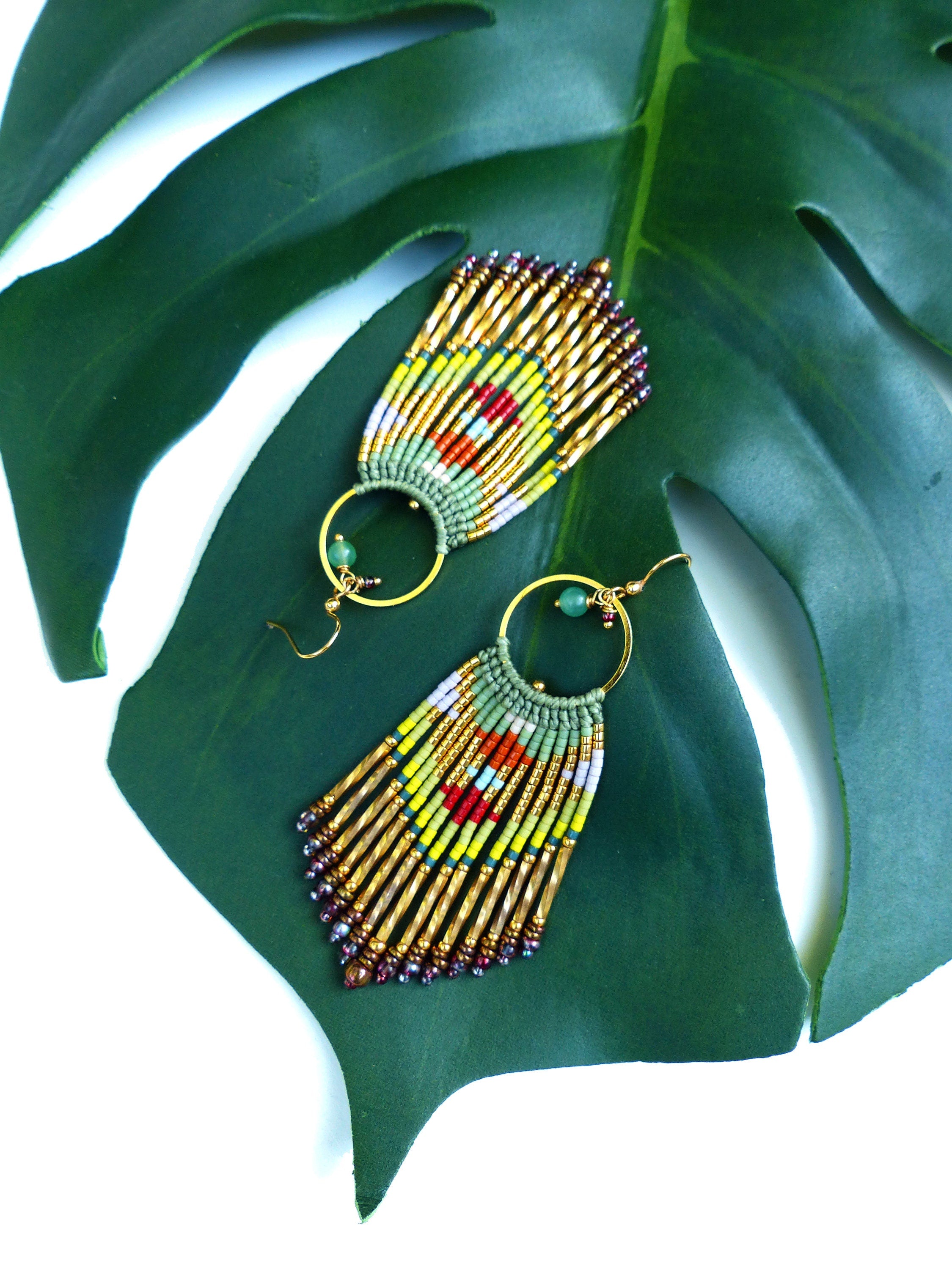 Peut inclure: Une paire de boucles d'oreilles créoles dorées avec une frange de perles. La frange est composée de perles vertes, jaunes, rouges et blanches. Les boucles d'oreilles sont suspendues à une feuille verte.