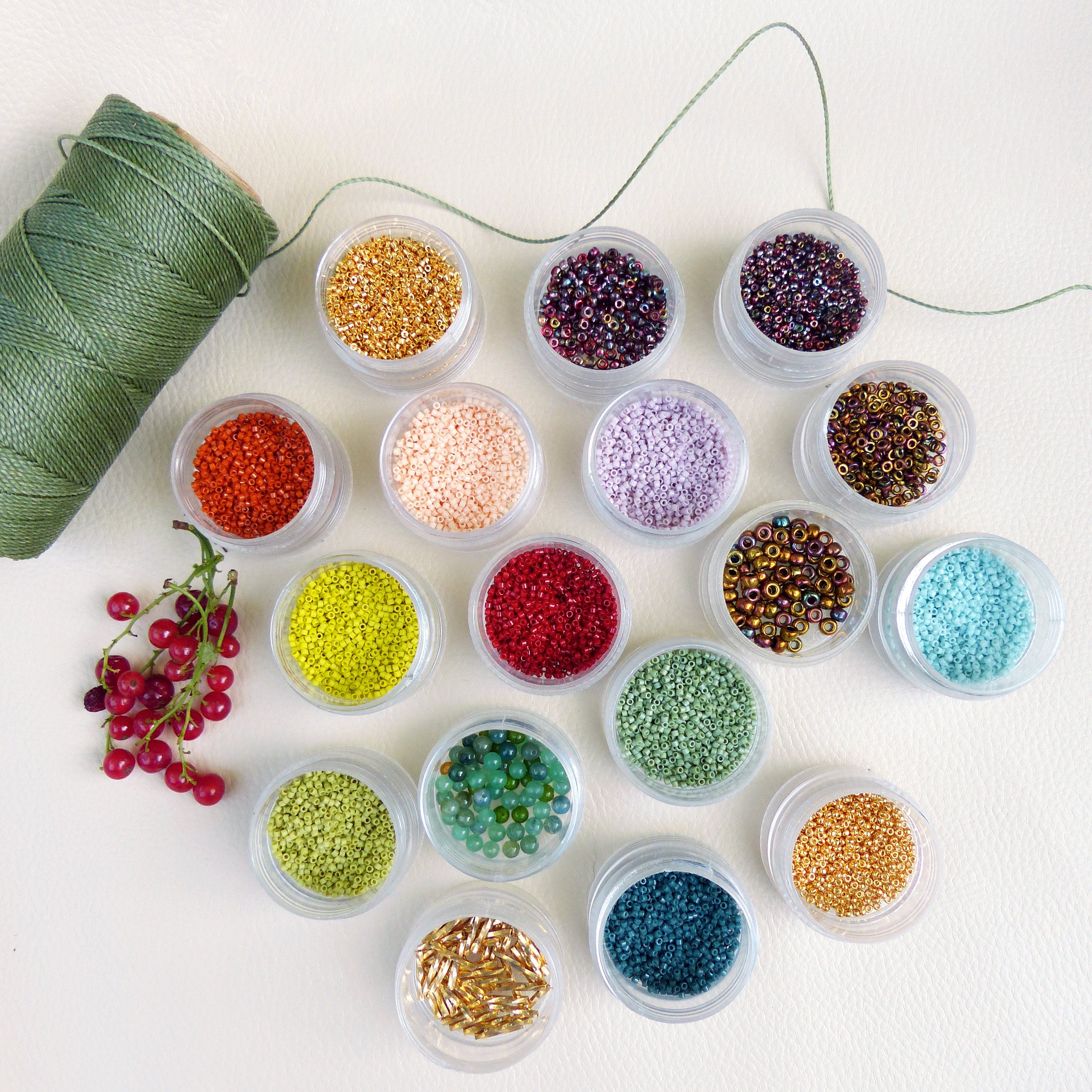Peut inclure: Assortiment de petits contenants remplis de perles colorées, disposés sur une surface blanche. Les contenants sont en verre transparent et présentent une variété de couleurs, notamment le rouge, l'orange, le jaune, le vert, le bleu, le violet et le marron. Il y a également une bobine de fil vert dans le coin supérieur gauche de l'image.