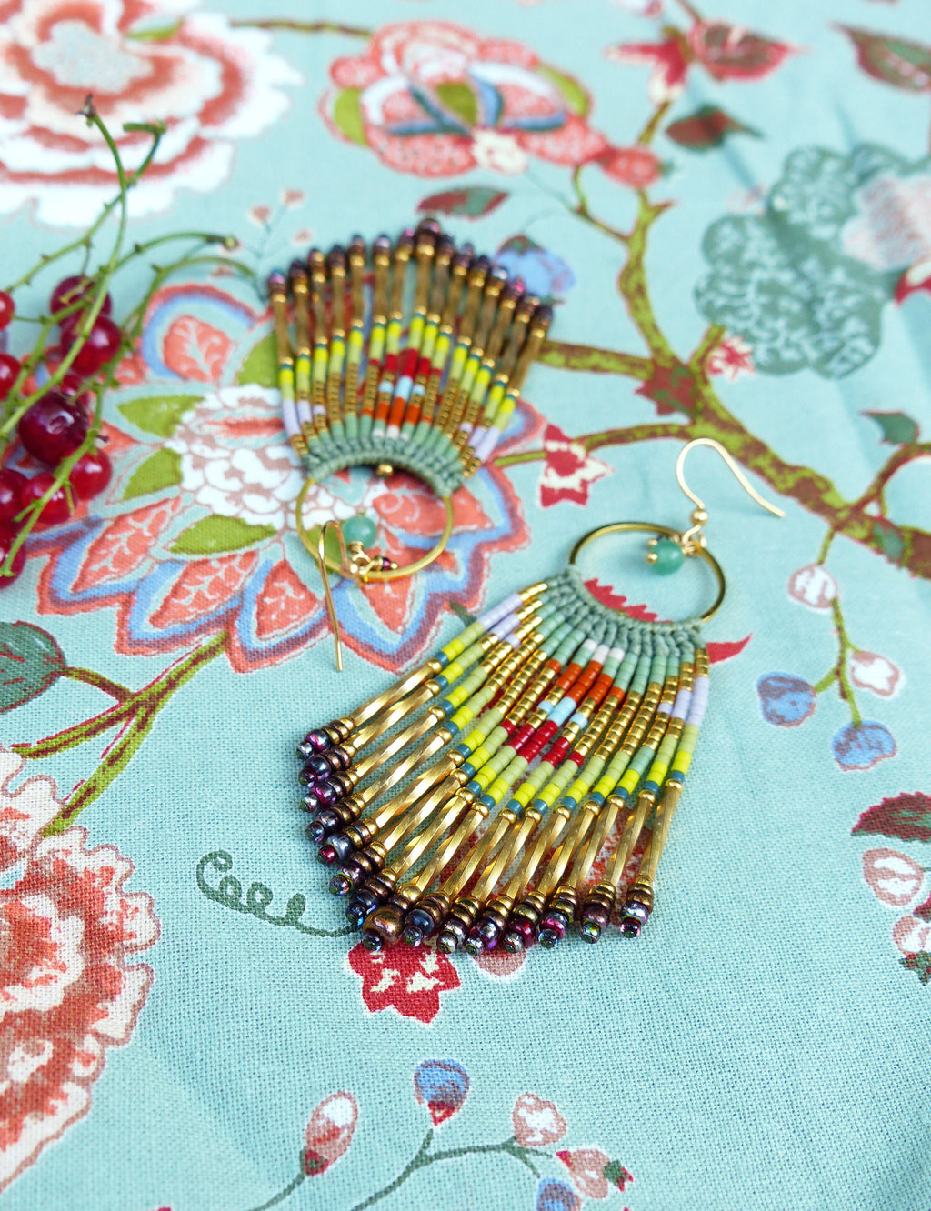 Peut inclure: Une paire de boucles d'oreilles créoles dorées avec un accent de pierre verte et une frange de perles colorées. Les perles sont disposées selon un motif géométrique et comprennent des nuances de vert, de jaune, de rouge et d'orange.