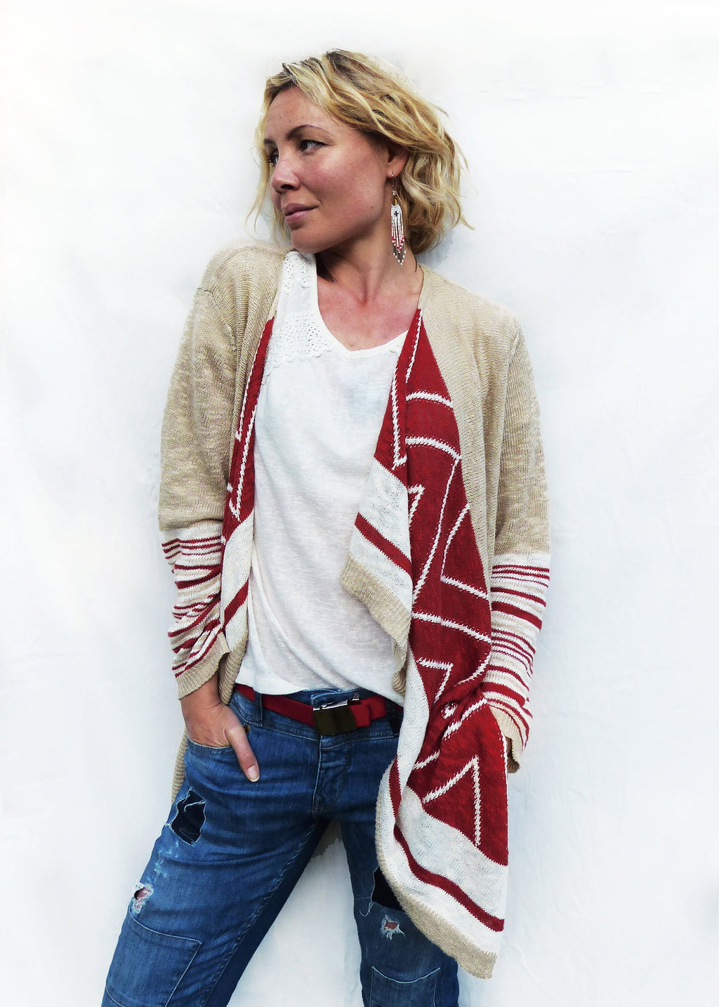 Peut inclure: Une femme porte un cardigan beige orné de motifs géométriques rouges et blancs. Elle est vêtue d'un haut blanc, d'un jean bleu et d'une ceinture rouge. L'ensemble est décontracté et élégant.