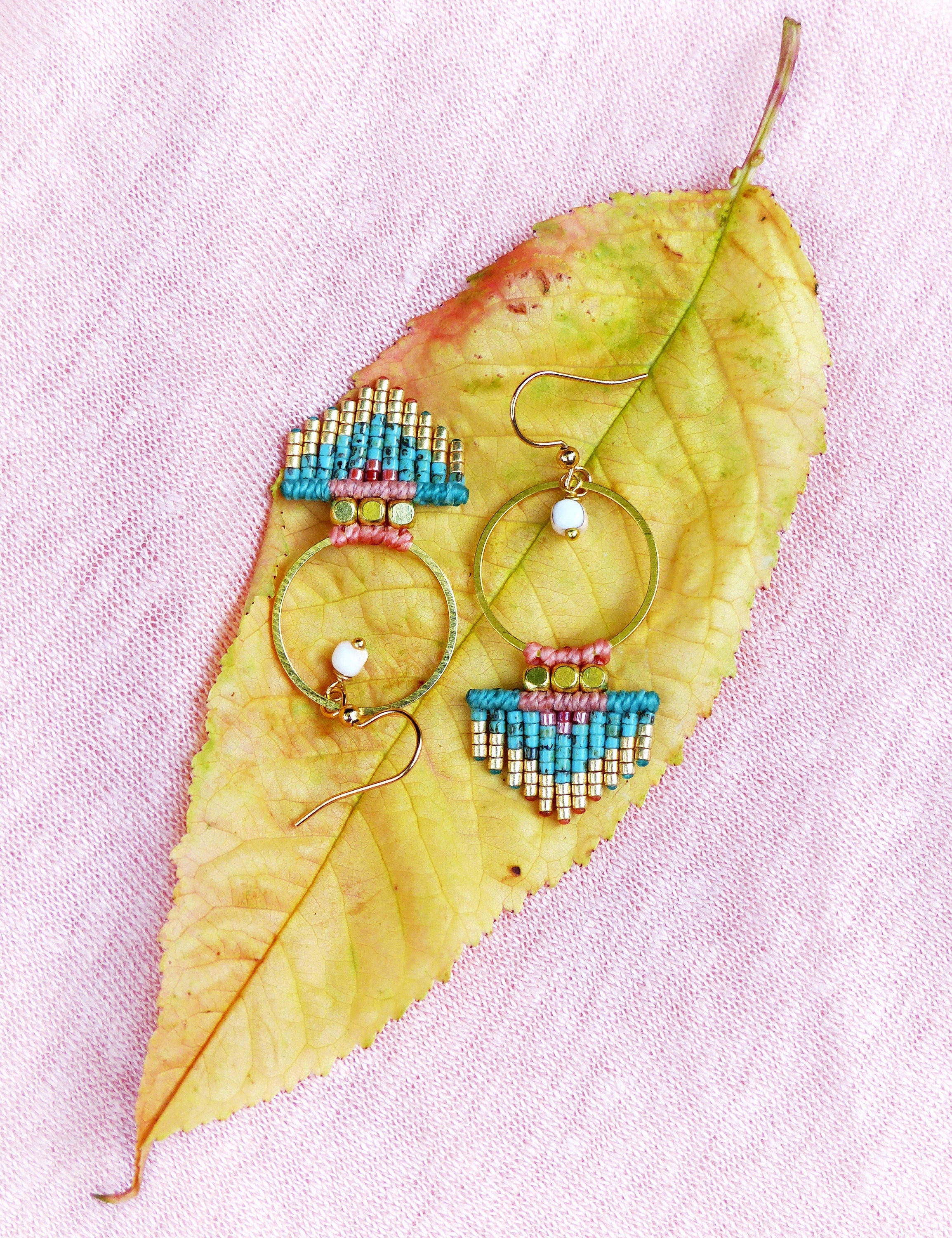 Peut inclure: Boucles d'oreilles artisanales avec des cercles dorés et des perles turquoise, dorées, roses et rouges. Elles sont posées sur une feuille jaune, sur un fond rose.