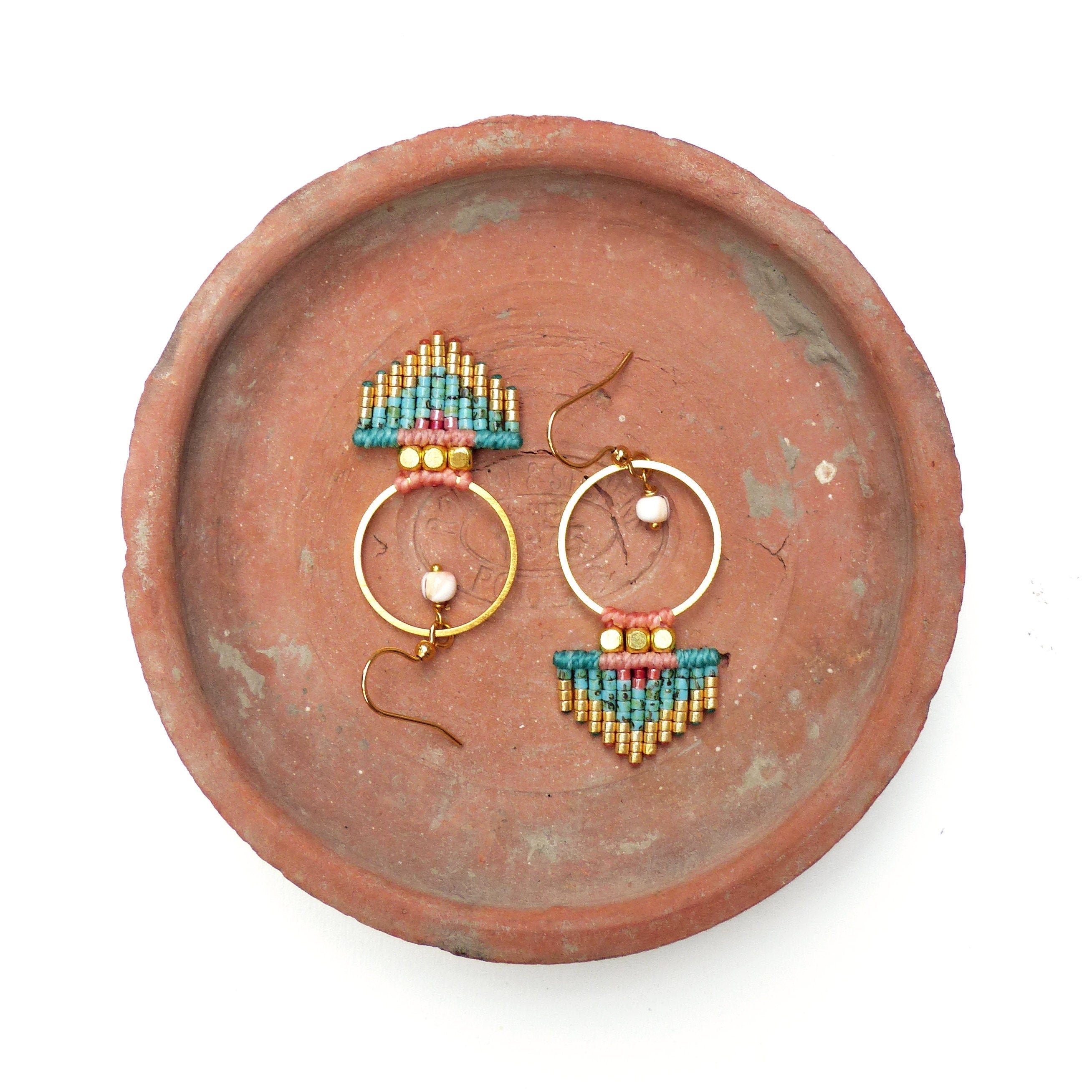 Peut inclure: Boucles d'oreilles faites à la main avec des cerceaux dorés et des détails en macramé. Les boucles d'oreilles présentent des perles turquoise, roses et dorées, avec une petite perle blanche au centre de chaque cercle. Présentées sur une assiette en terre cuite.