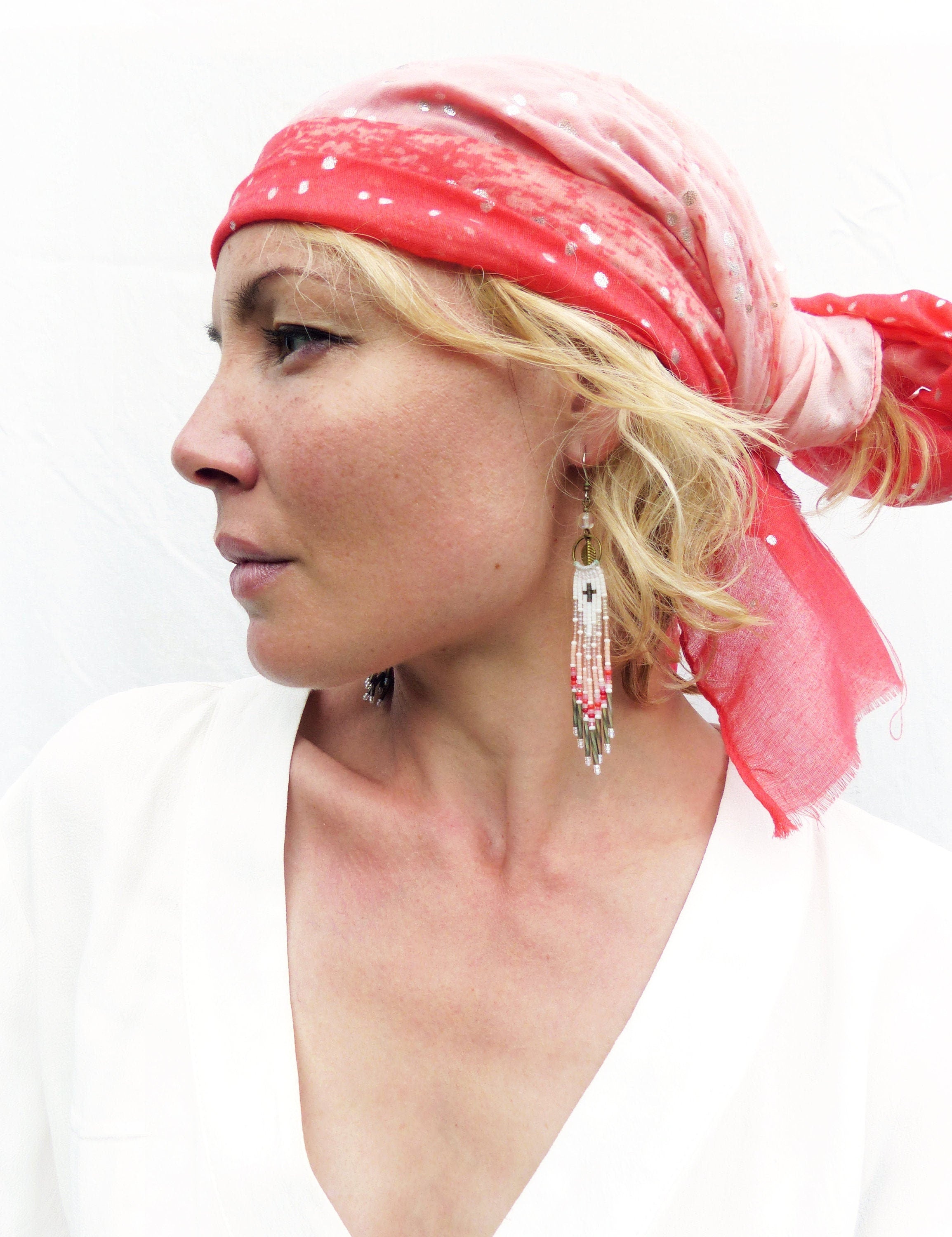 Peut inclure: Une femme porte un foulard rouge et rose à pois blancs et un chemisier blanc. La femme porte de longues boucles d'oreilles perlées avec un motif de croix et des franges.
