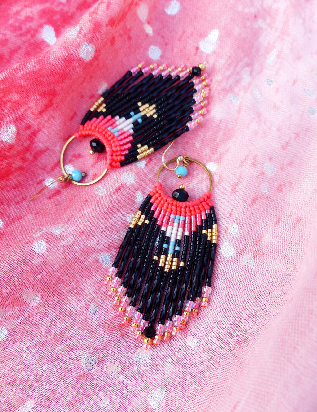 Peut inclure: Une paire de longues boucles d'oreilles en perles avec un motif géométrique en noir, rose, or et blanc. Les boucles d'oreilles sont suspendues à des créoles dorées.