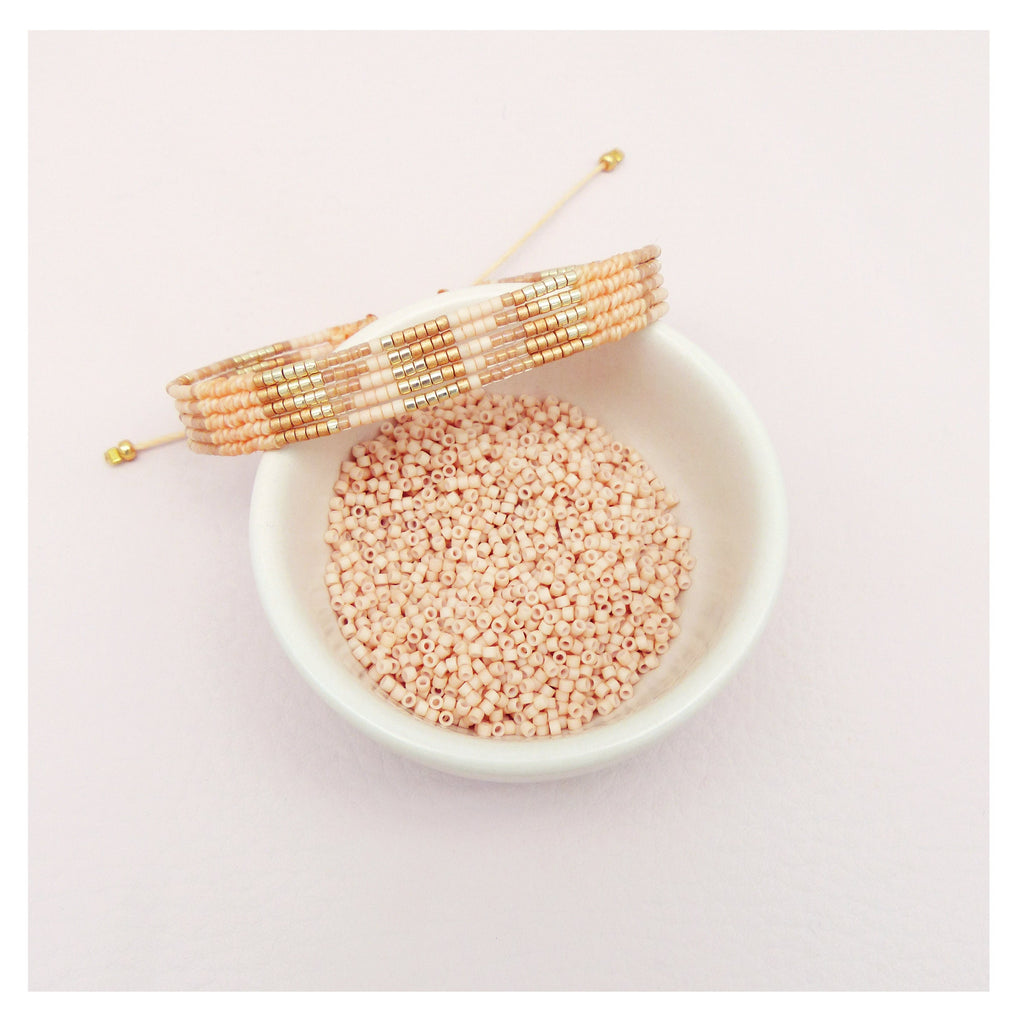 Peut inclure: Un gros plan d'un bol blanc rempli de petites perles rose pâle. Un bracelet en perles avec des perles dorées et blanches est posé sur le bol.