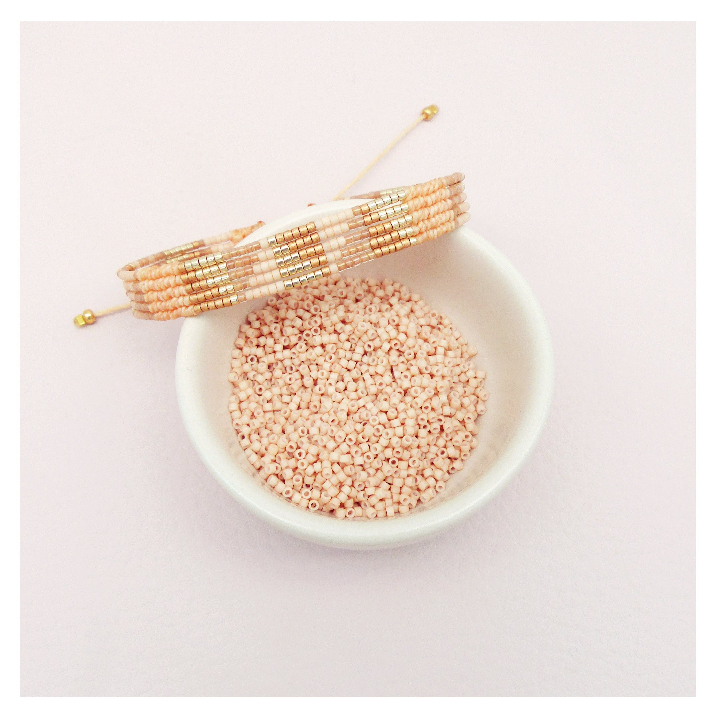 Peut inclure: Un gros plan d'un bol blanc rempli de petites perles rose pâle. Un bracelet en perles avec des perles dorées et blanches est posé sur le bol.