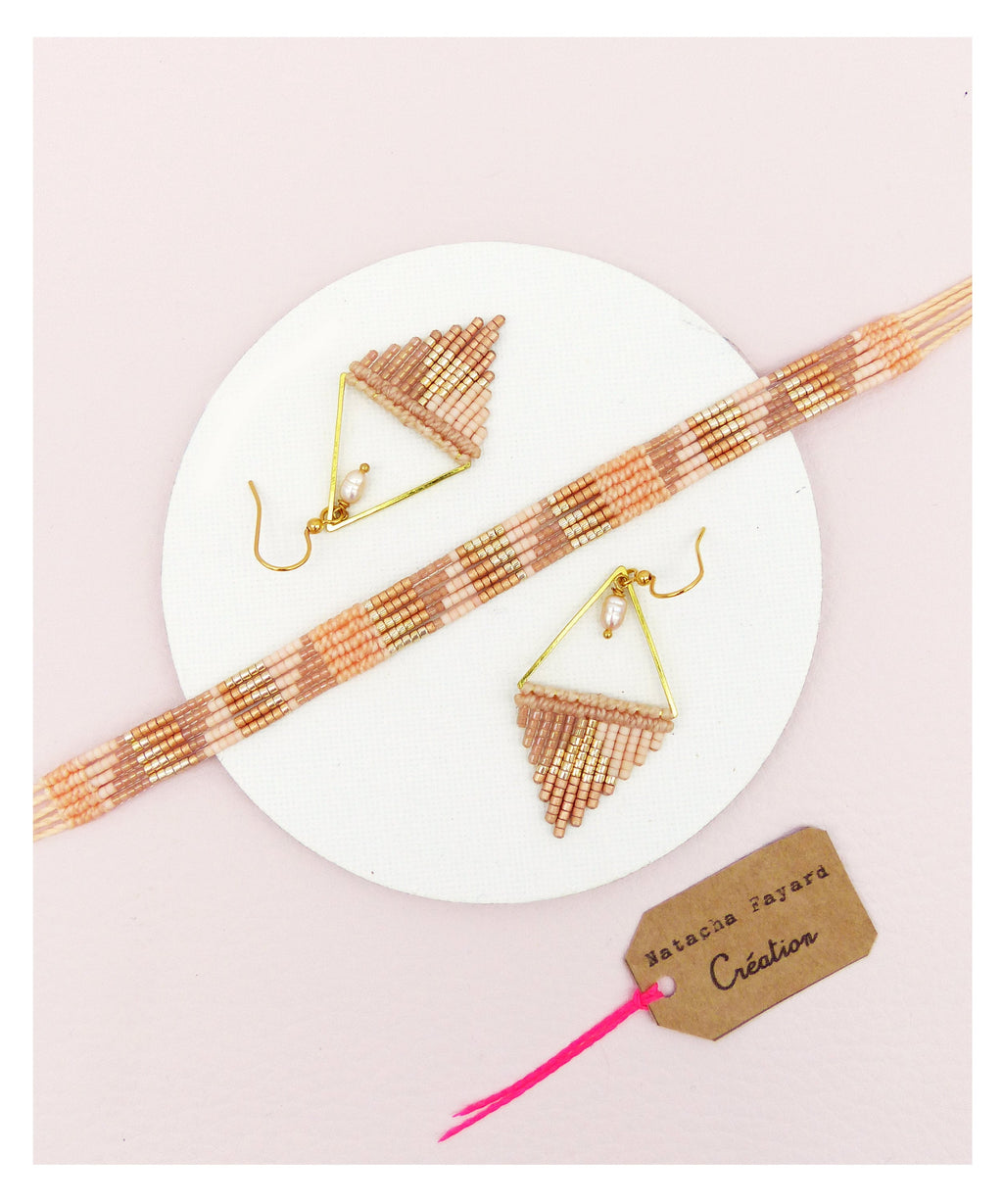 Peut inclure: Un ensemble de bijoux en perles roses et dorées comprenant une paire de boucles d'oreilles triangulaires et un bracelet. Les boucles d'oreilles sont ornées d'une petite perle. Le bracelet et les boucles d'oreilles sont faits de petites perles colorées. Les bijoux sont présentés sur une assiette blanche avec une étiquette brune sur laquelle est inscrit "Natacha Zayard Création".