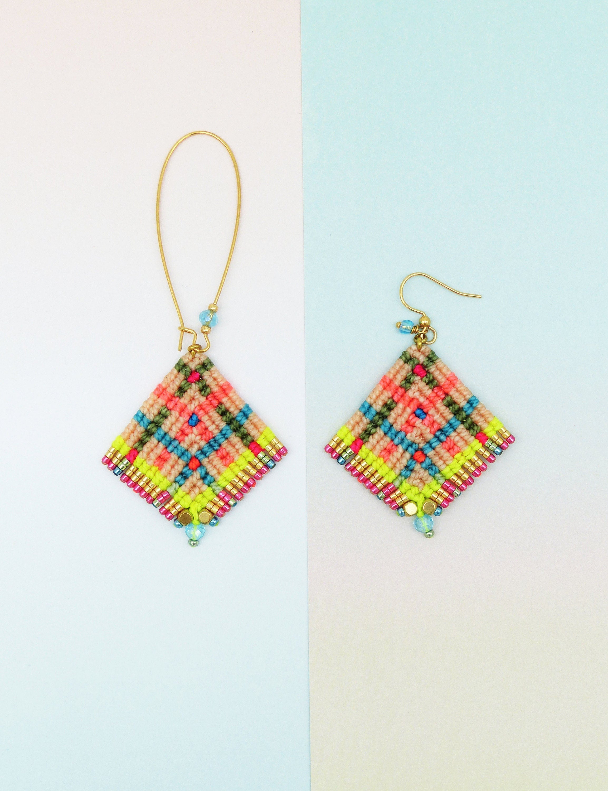 Peut inclure: Deux boucles d'oreilles faites à la main avec un motif géométrique. Les boucles d'oreilles ont une forme de losange avec une base beige, accentuée de fils rose fluo, verts et bleus. Des crochets dorés et de petites perles complètent le design. Sur fond blanc et bleu clair.
