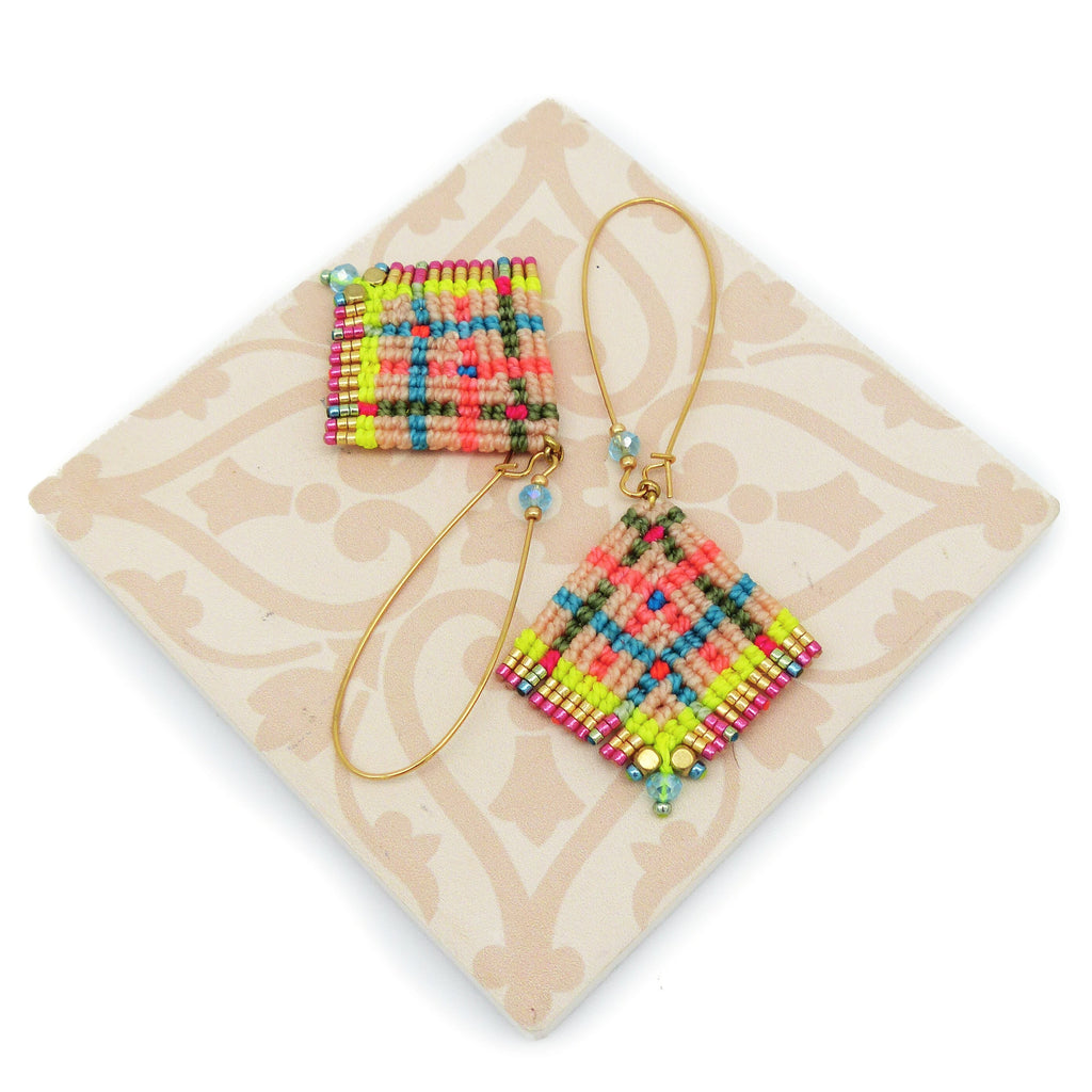 Peut inclure: Une paire de boucles d'oreilles macramé colorées. Les boucles d'oreilles présentent une forme carrée et une forme de losange, avec un motif tissé en rose, bleu, vert et jaune. Les boucles d'oreilles sont attachées à des crochets d'oreilles dorés et ornées de petites perles.