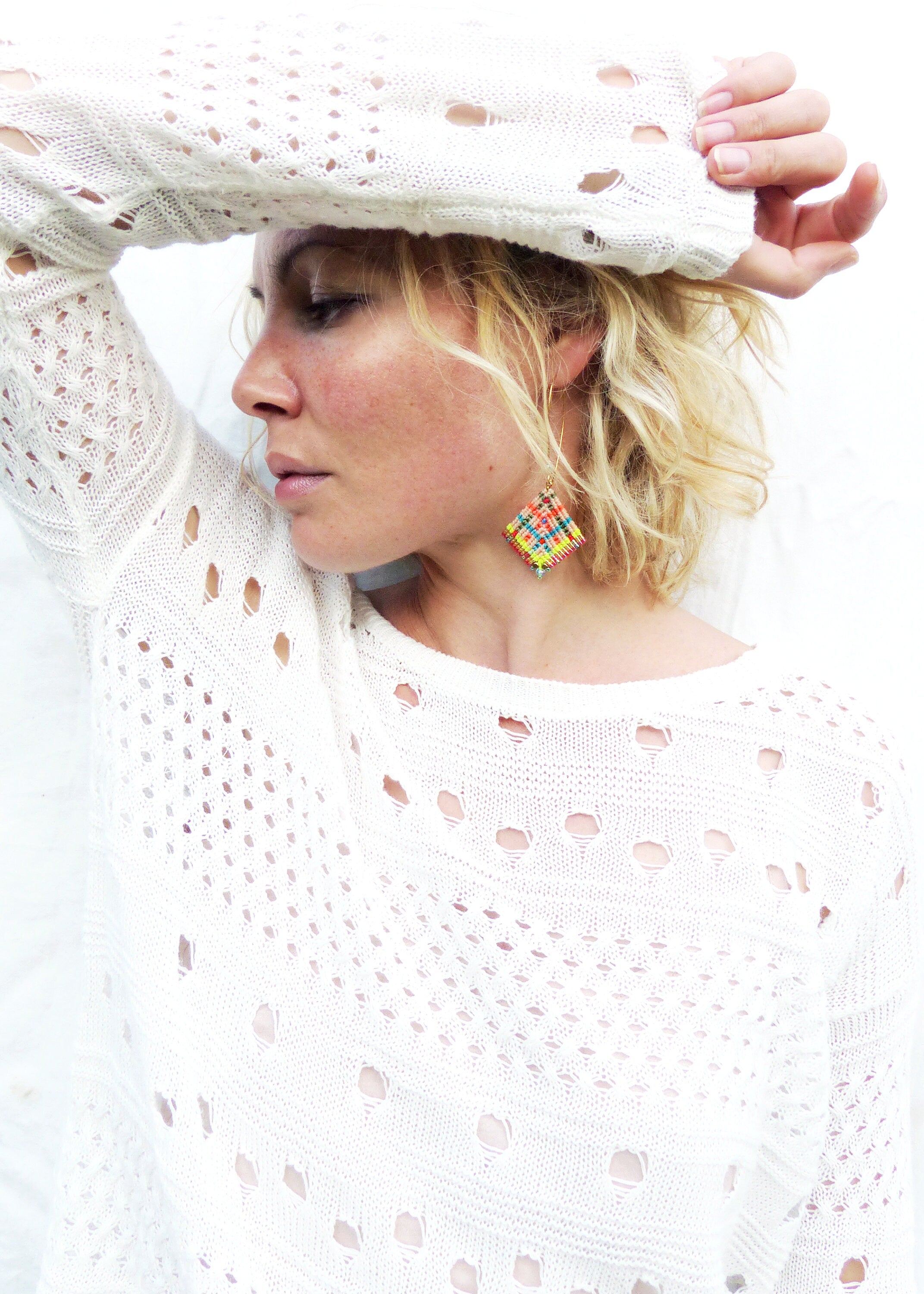 Peut inclure: Une femme porte un pull blanc en tricot ajouré avec une boucle d'oreille en forme de losange. La boucle d'oreille présente un motif tissé coloré de fils jaunes, rouges, bleus et verts. Le pull a un col rond et des manches longues.