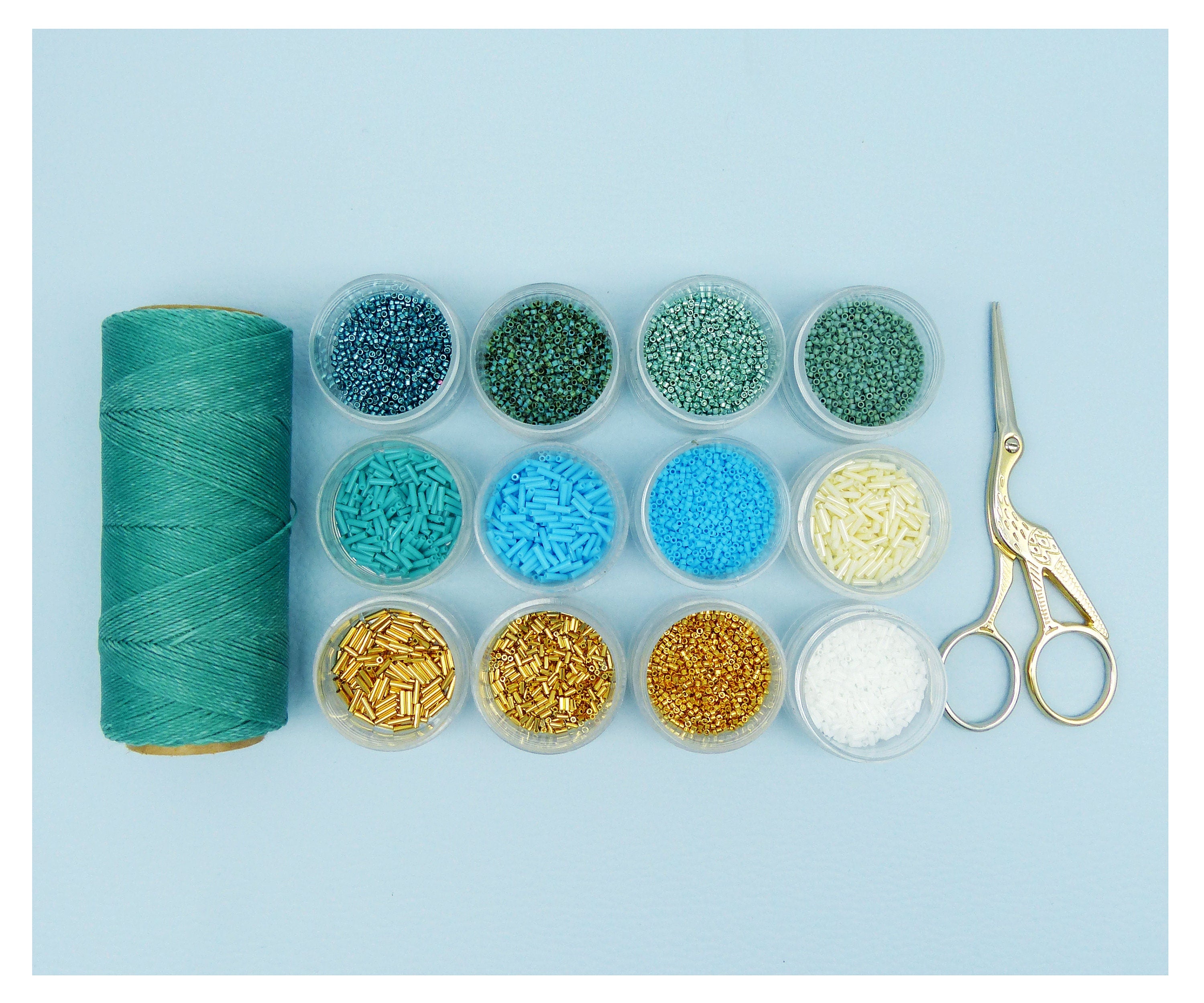 Peut inclure: Un écheveau de fil vert-bleu, de petits récipients de perles colorées et des ciseaux dorés avec un motif d'oiseau sont disposés sur une surface bleu clair. Les perles sont de différentes nuances de bleu, vert, or et blanc. Les ciseaux mesurent environ 10 cm.