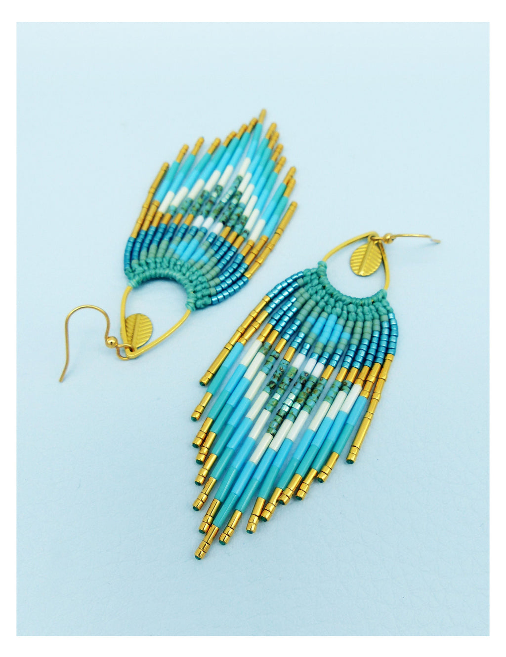 Peut inclure: Une paire de boucles d'oreilles en perles avec un motif de plumes. Les boucles d'oreilles présentent des perles turquoise, blanches et dorées. La partie supérieure est de couleur sarcelle et les boucles d'oreilles ont des crochets dorés.