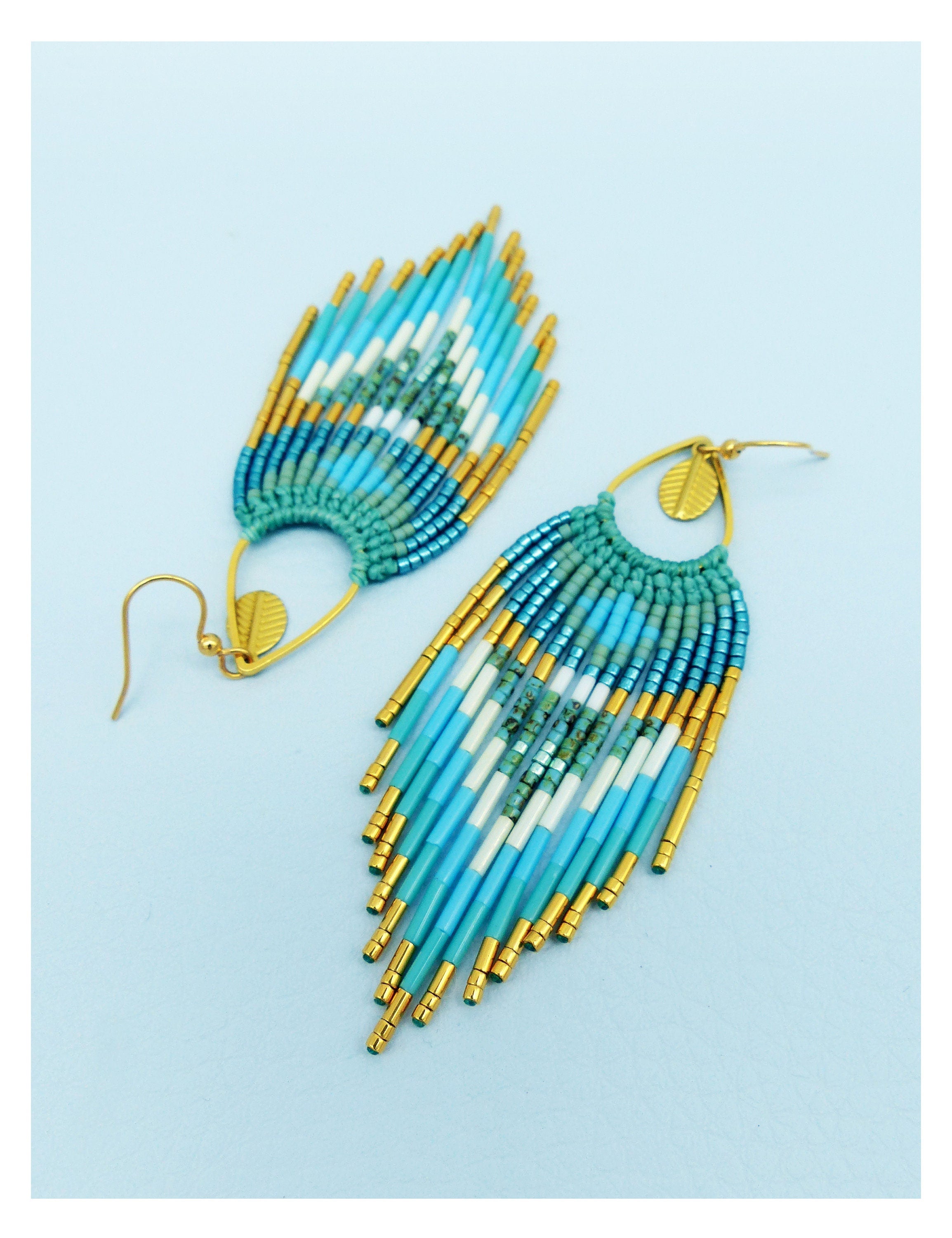 Peut inclure: Une paire de boucles d'oreilles en perles avec un motif de plumes. Les boucles d'oreilles présentent des perles turquoise, blanches et dorées. La partie supérieure est de couleur sarcelle et les boucles d'oreilles ont des crochets dorés.