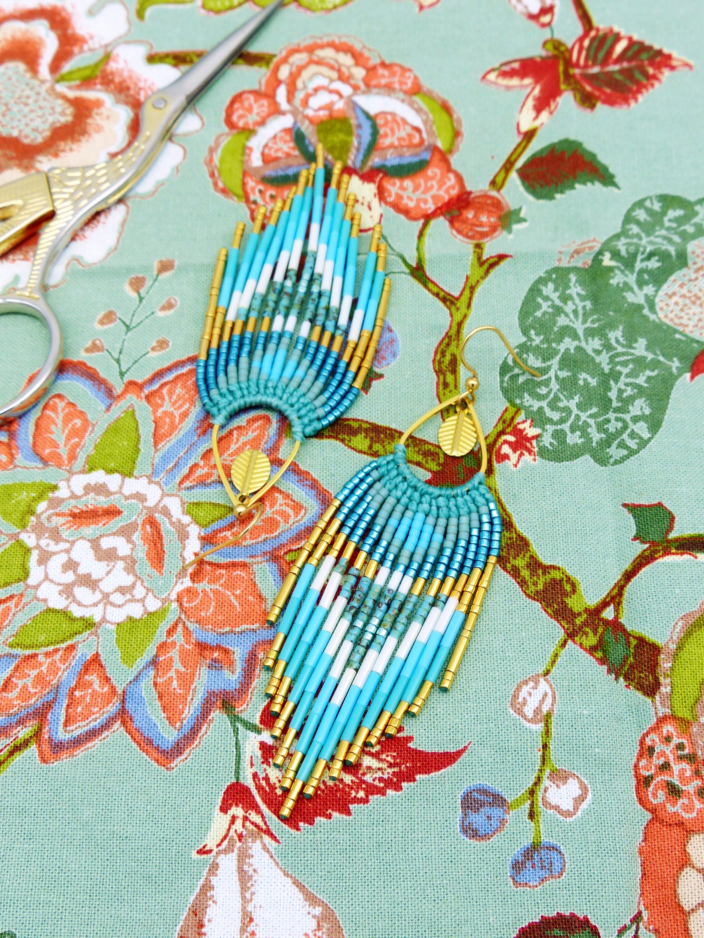 Peut inclure: Une paire de boucles d'oreilles à franges en perles turquoise, blanches et dorées, avec des accents dorés en forme de feuille. Les boucles d'oreilles sont présentées sur un tissu à motifs floraux, avec des ciseaux en arrière-plan.