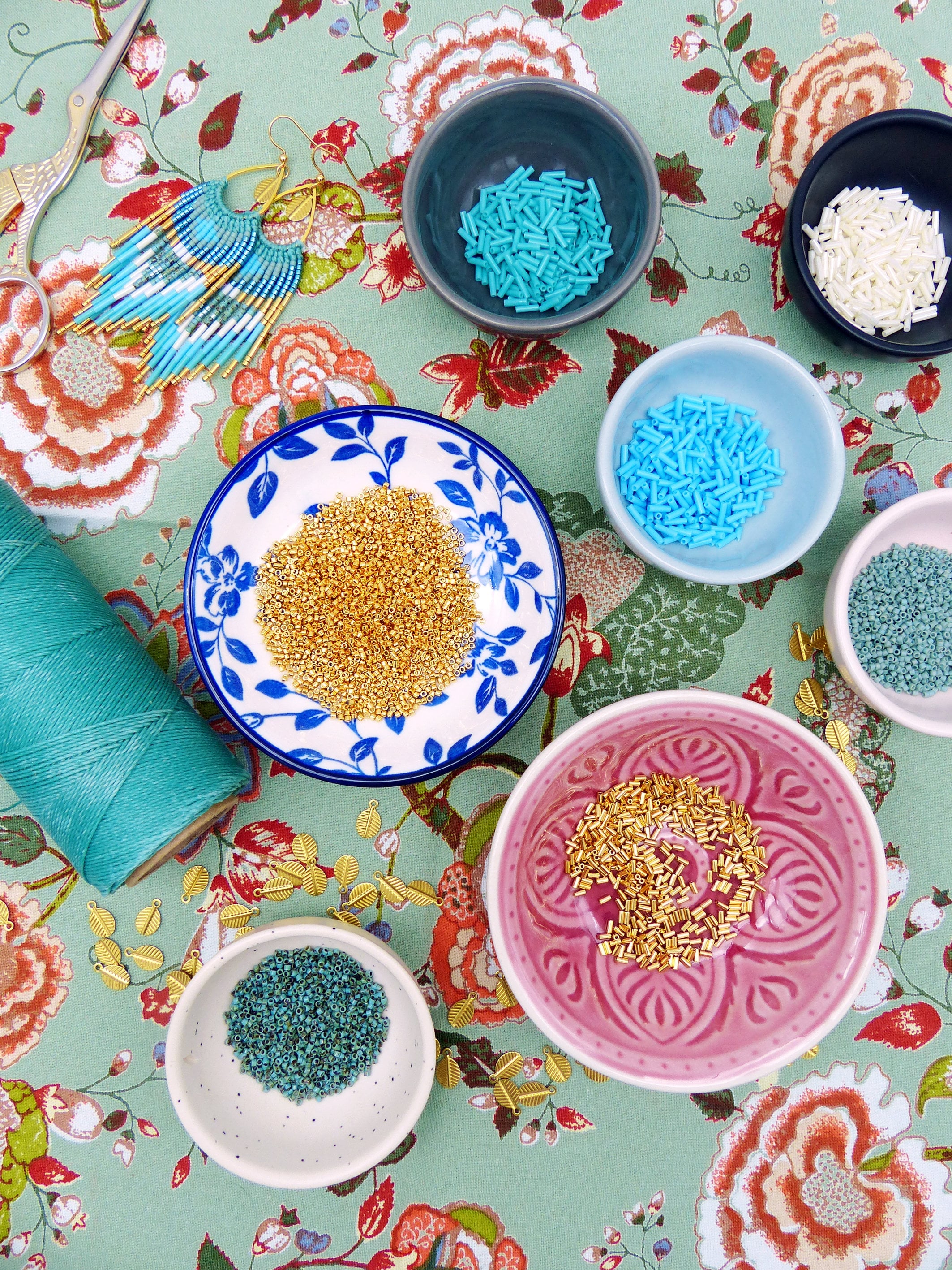 Peut inclure: Composition de fournitures de création de bijoux sur un tissu à motifs floraux. Petits bols remplis de perles de différentes couleurs, une bobine de fil turquoise et des ciseaux. Une paire de boucles d'oreilles en perles turquoise et dorées est également présente.