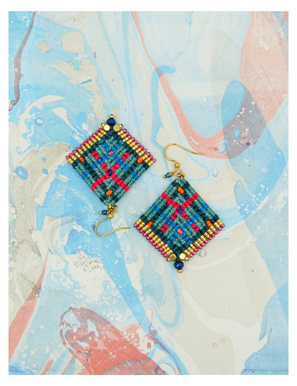 Peut inclure: Une paire de boucles d'oreilles macramé faites à la main. Chaque boucle d'oreille présente un motif en forme de losange avec un motif tissé turquoise, rouge et bleu, orné de perles dorées et d'un crochet doré. Les boucles d'oreilles sont sur un fond marbré.
