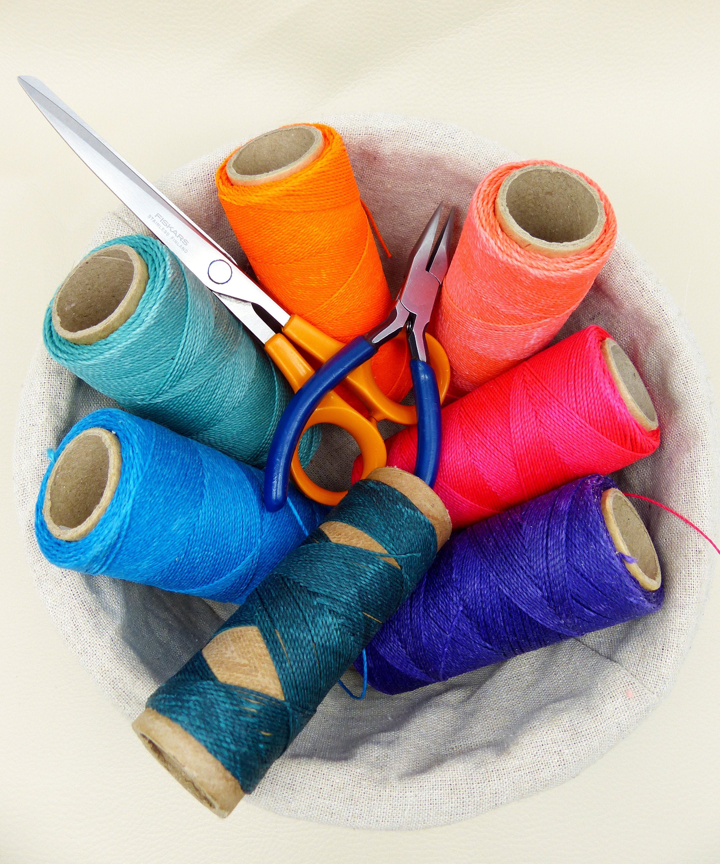 Peut inclure: Un panier rempli de bobines de fil colorées, dans des tons orange, rose, bleu, turquoise et violet. Une paire de ciseaux à poignée orange et une pince à poignée bleue suggèrent un thème de couture ou de bricolage.