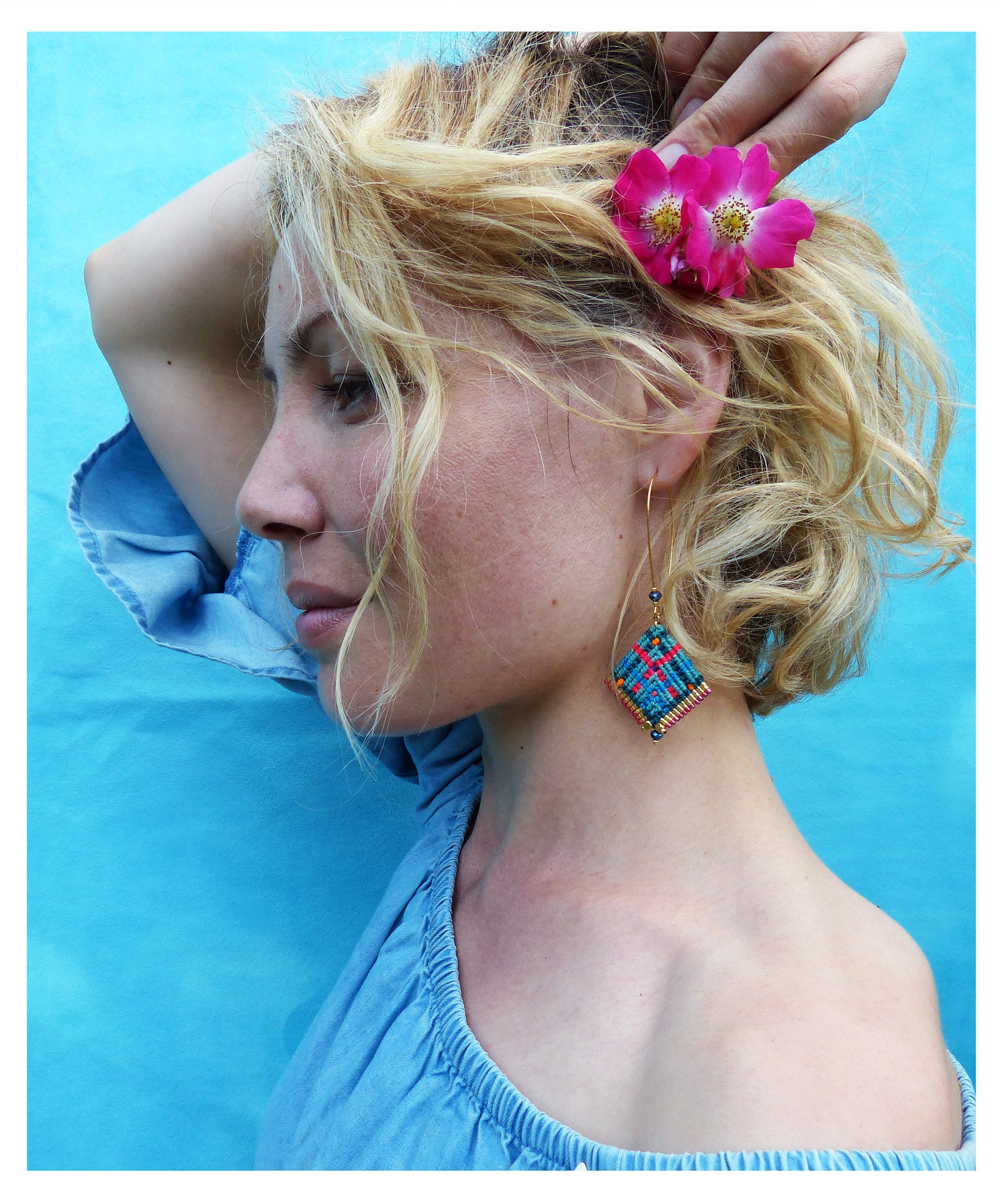 Peut inclure: Une femme aux cheveux blonds porte une fleur rose dans les cheveux, un haut bleu à épaules dénudées et des boucles d'oreilles en macramé colorées. Les boucles d'oreilles sont en forme de losange avec des fils bleus, rouges et oranges.